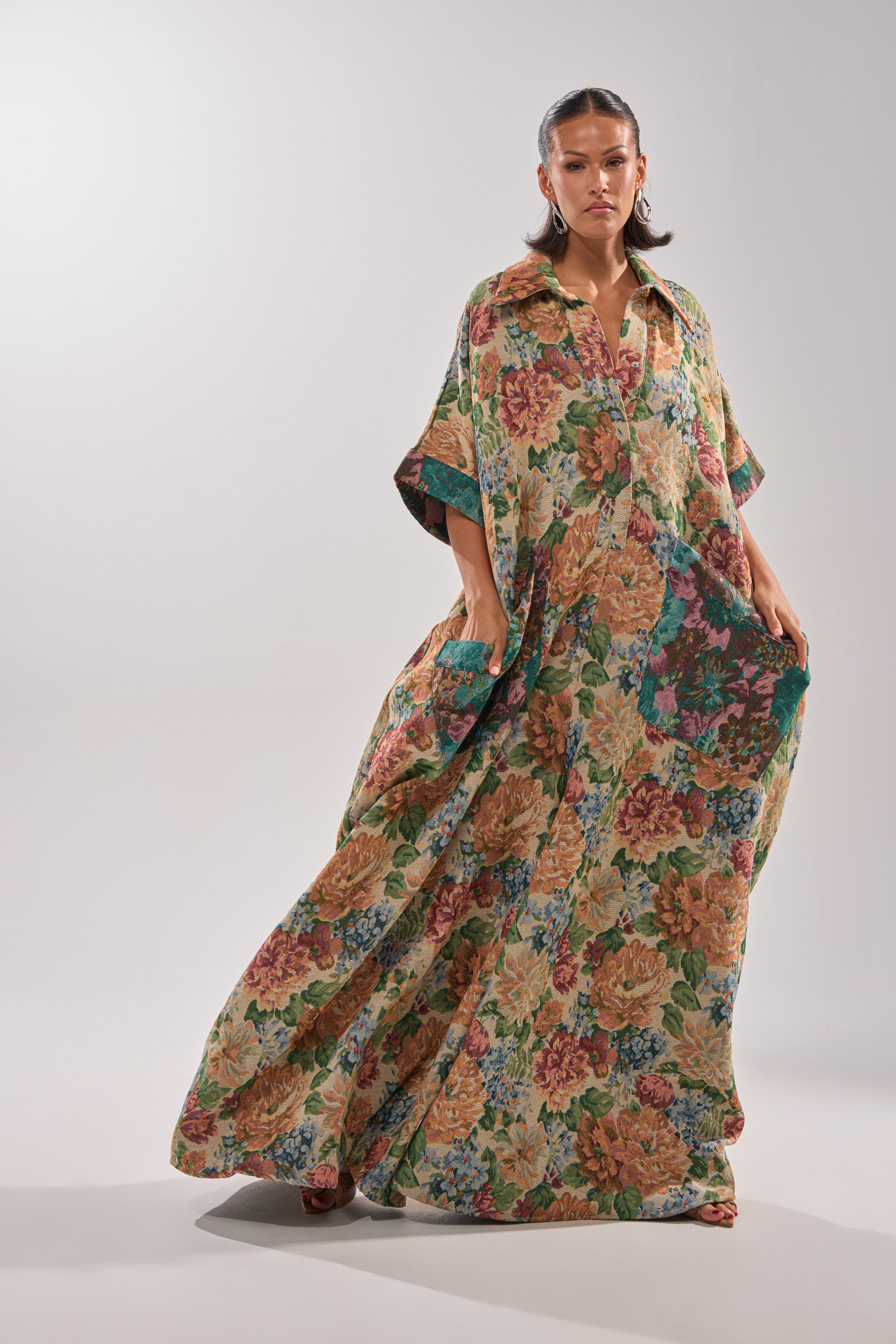 A woman stands in a studio wearing the VANJA BROCADE JUMPSUIT, featuring vintage-inspired tapestry fabric with a colorful floral pattern, short sleeves, pockets, and a loose fit. Her hair is styled and she has a serious expression.