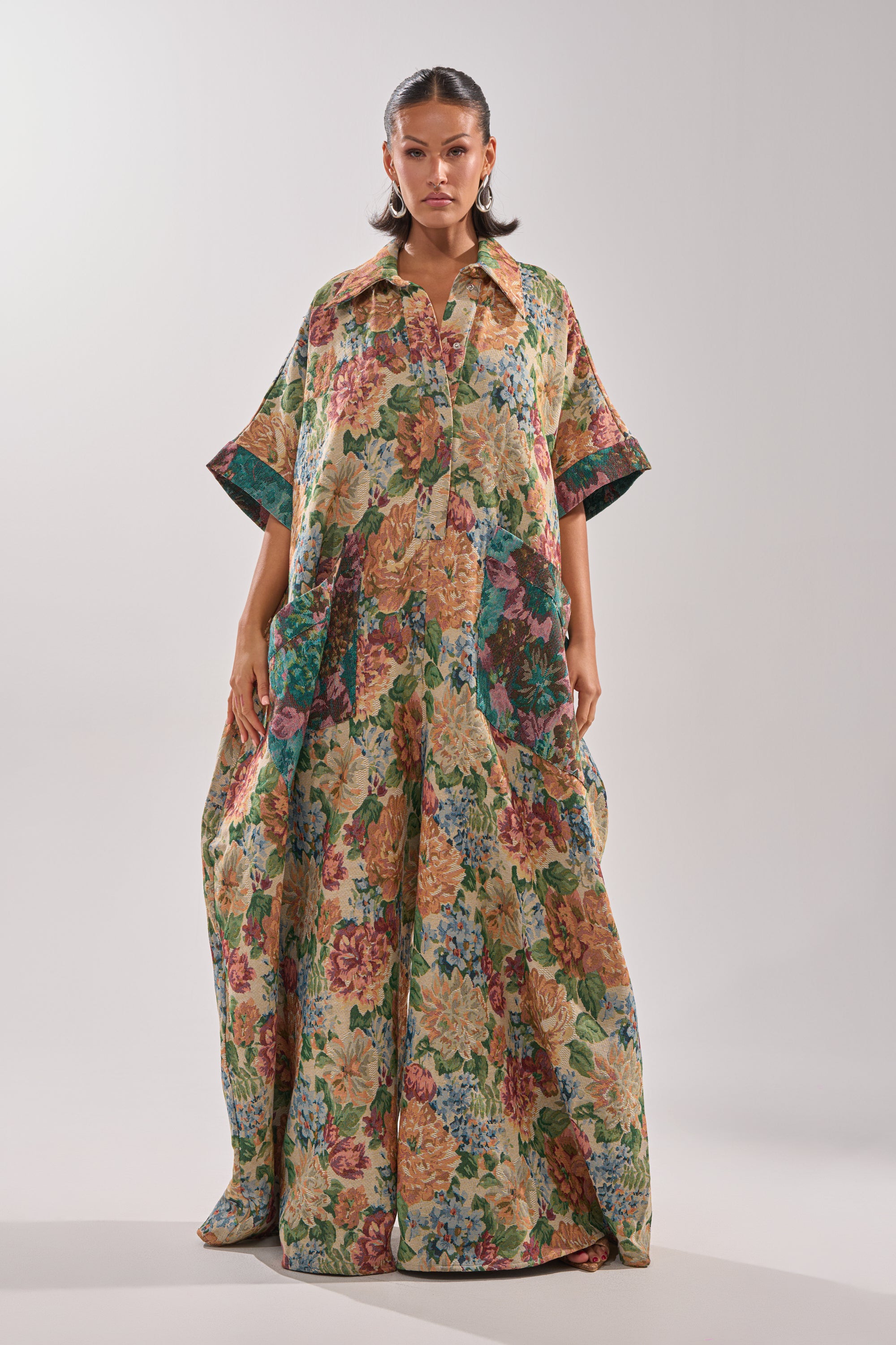 A woman faces forward in the VANJA BROCADE JUMPSUIT, featuring a loose, flowing fit, vibrant tapestry-inspired floral print, short sleeves, a collar, and front buttons. She stands against a plain light background.