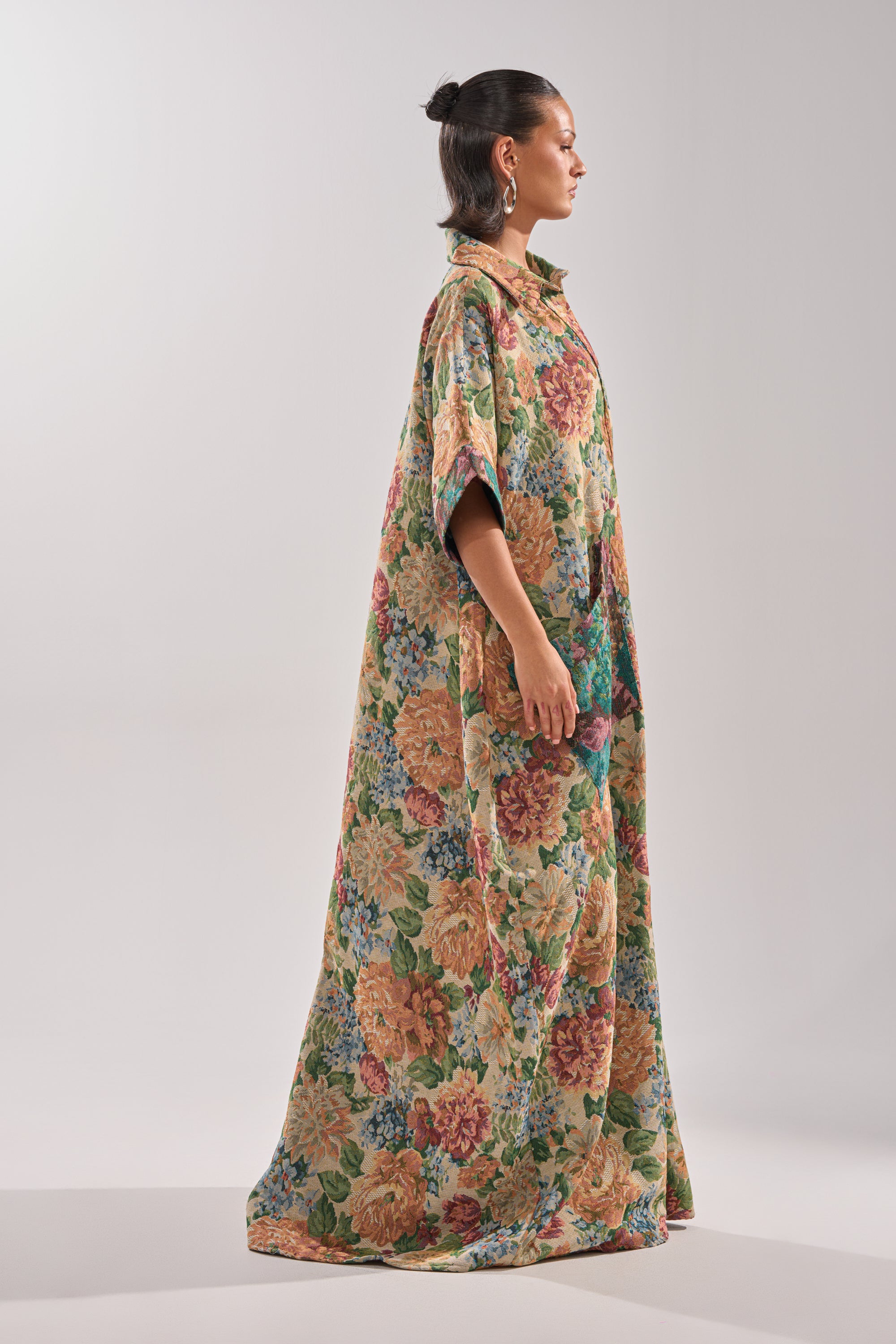 A person stands in profile wearing the VANJA BROCADE JUMPSUIT, a long, loose piece with wide sleeves and tapestry-style fabric. A plain background highlights its colorful, vintage-inspired pattern.