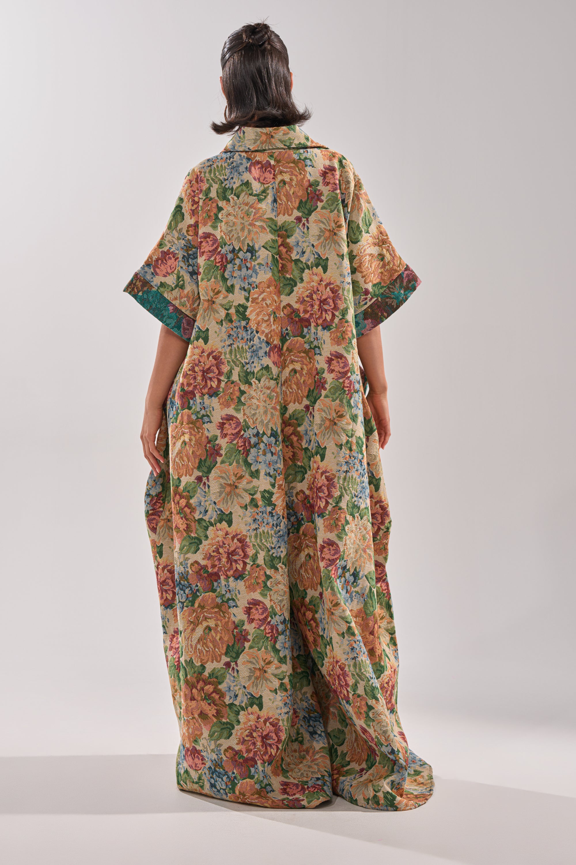 A person stands facing away, wearing the VANJA BROCADE JUMPSUIT—a long, loose garment with large colorful floral patterns and greenery on a light background. It features wide sleeves and floor-length draping fabric against a plain, light backdrop.