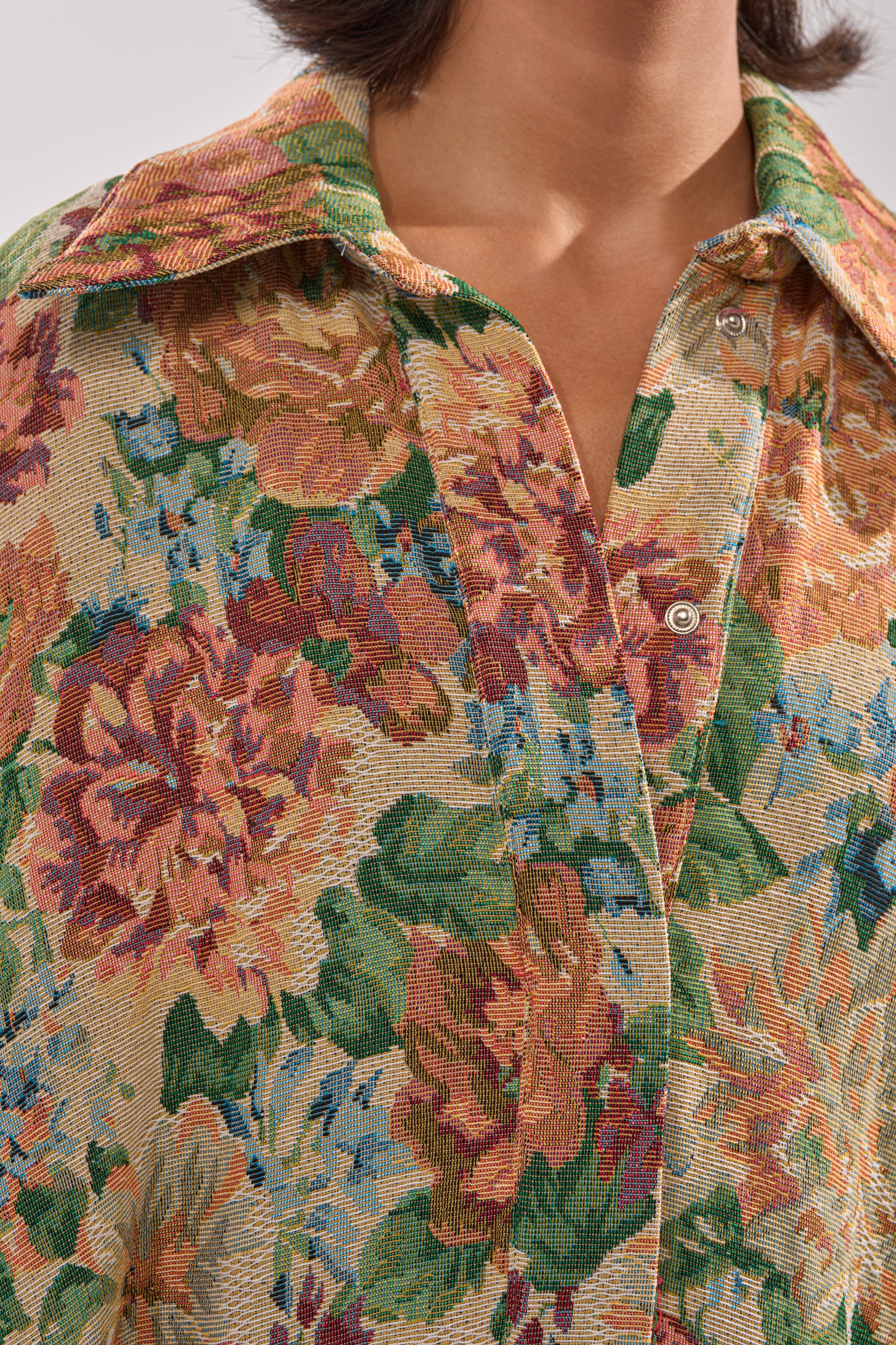 A close-up of a person wearing the VANJA BROCADE JUMPSUIT, made from tapestry-style fabric in green, pink, orange, and blue with buttons and textured details. The collared jumpsuit is shown without the person's face visible.