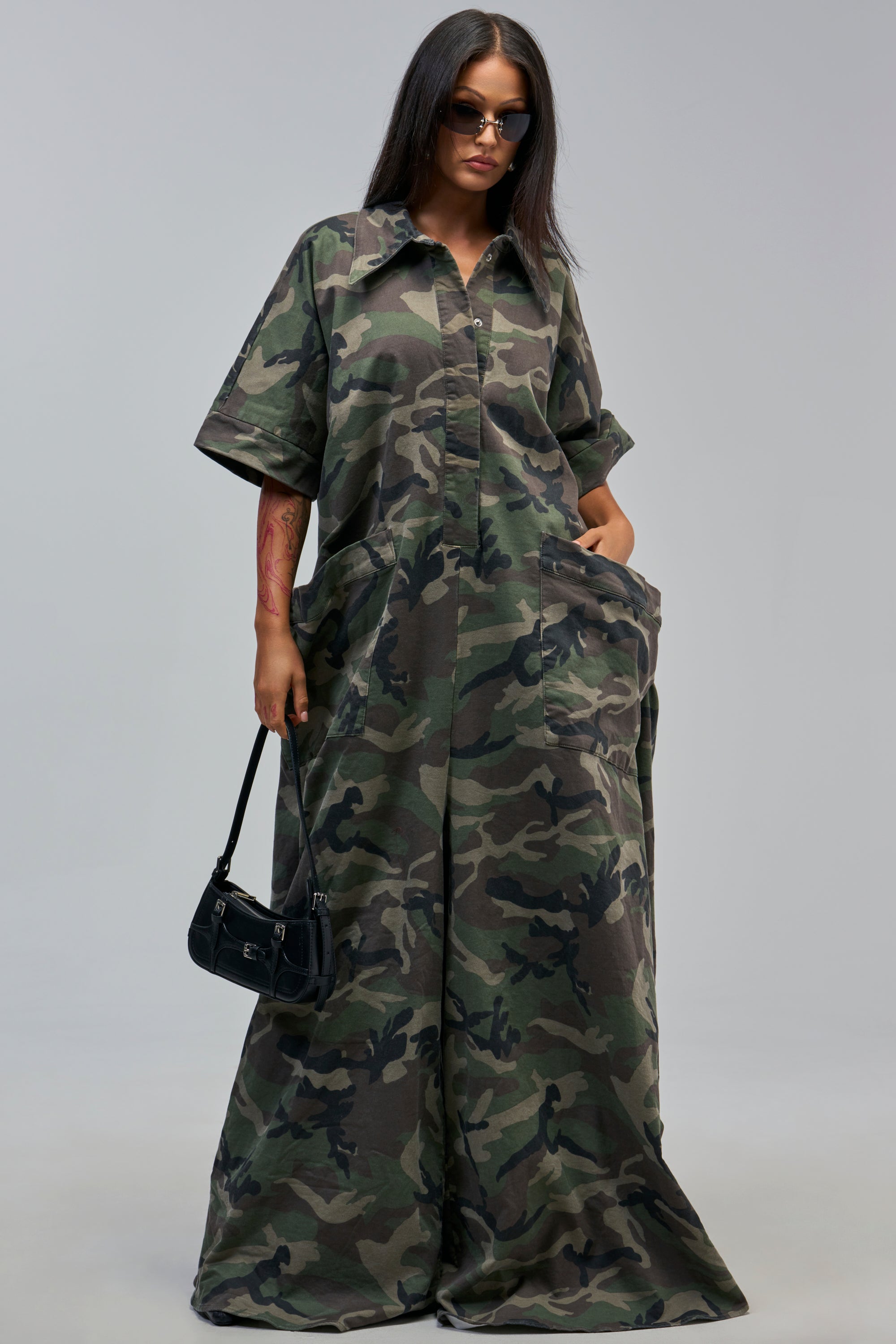 A woman in the SHE’S RICH OVERSIZED CAMO JUMPSUIT, sunglasses, and a small black handbag stands against a plain gray background.