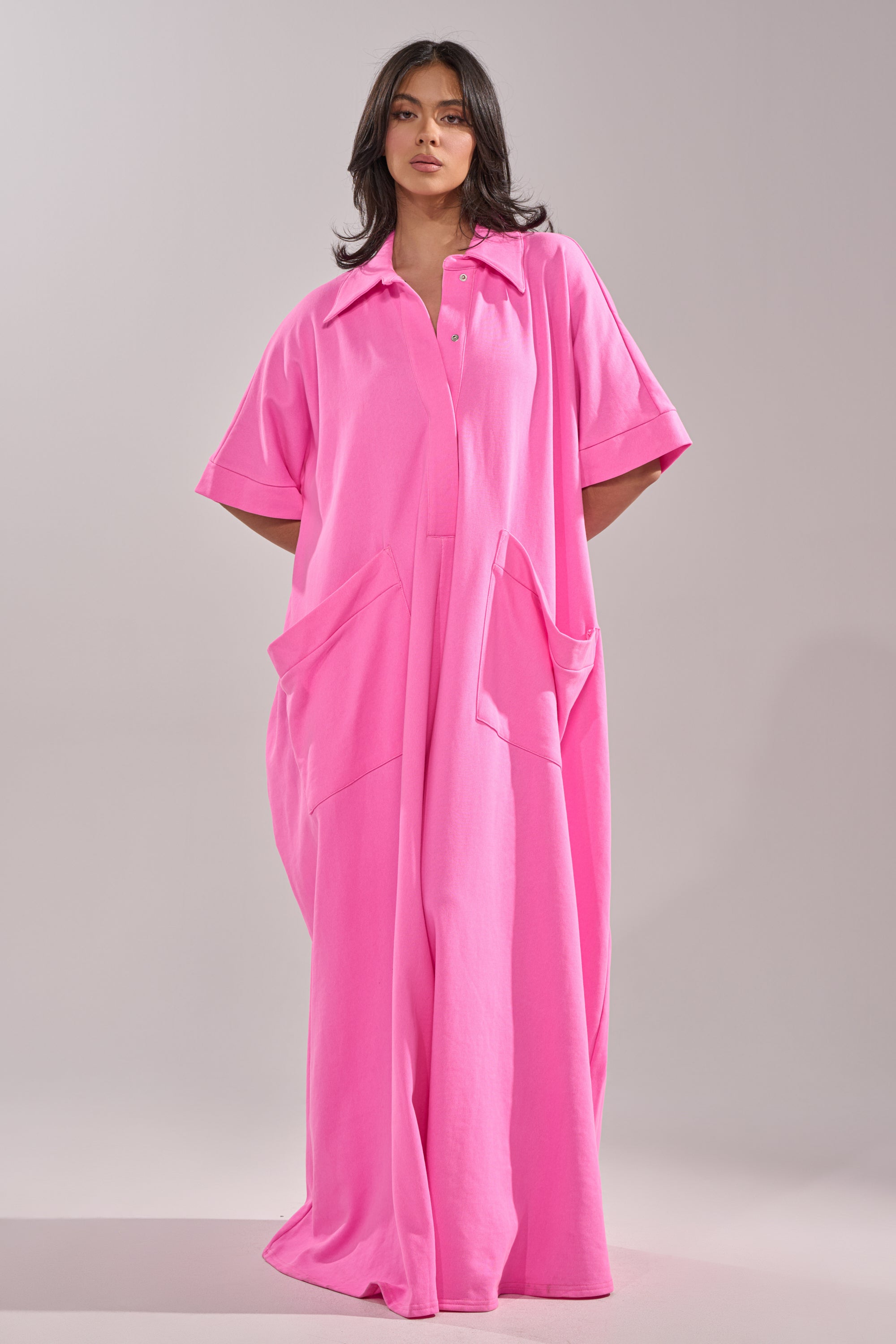 A woman with medium-length dark hair and a neutral expression models the PRETTY IN PINK JUMPSUIT, an oversized bright pink one-piece featuring short sleeves, a collar, and large front pockets, embodying effortless resort wear style.