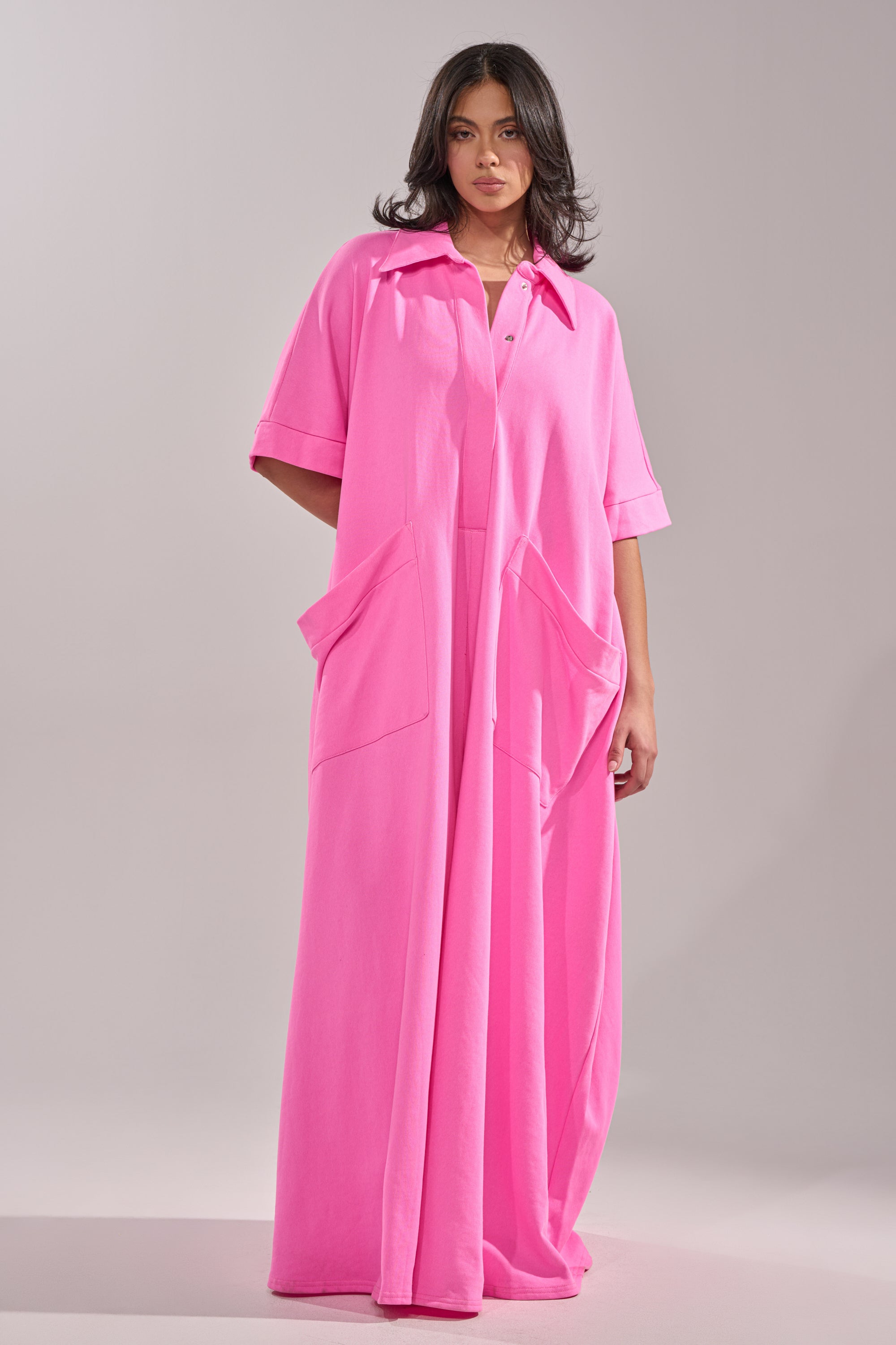 A woman wears the PRETTY IN PINK JUMPSUIT, an oversized bright pink one-piece with short sleeves, large front pockets, and a collar, standing against a plain light background.