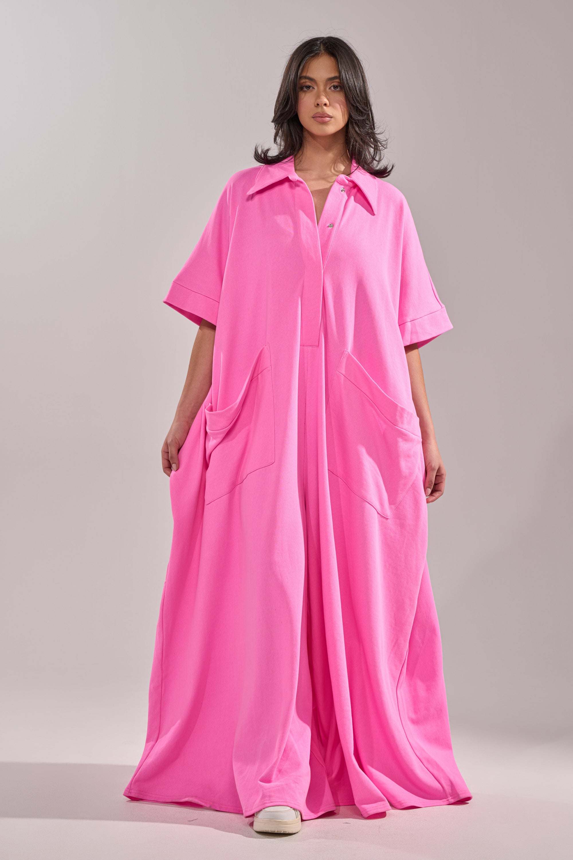 A woman faces forward in the PRETTY IN PINK JUMPSUIT, a bright pink, wide-leg style with short sleeves and large front pockets. She pairs it with white shoes against a plain light gray backdrop.