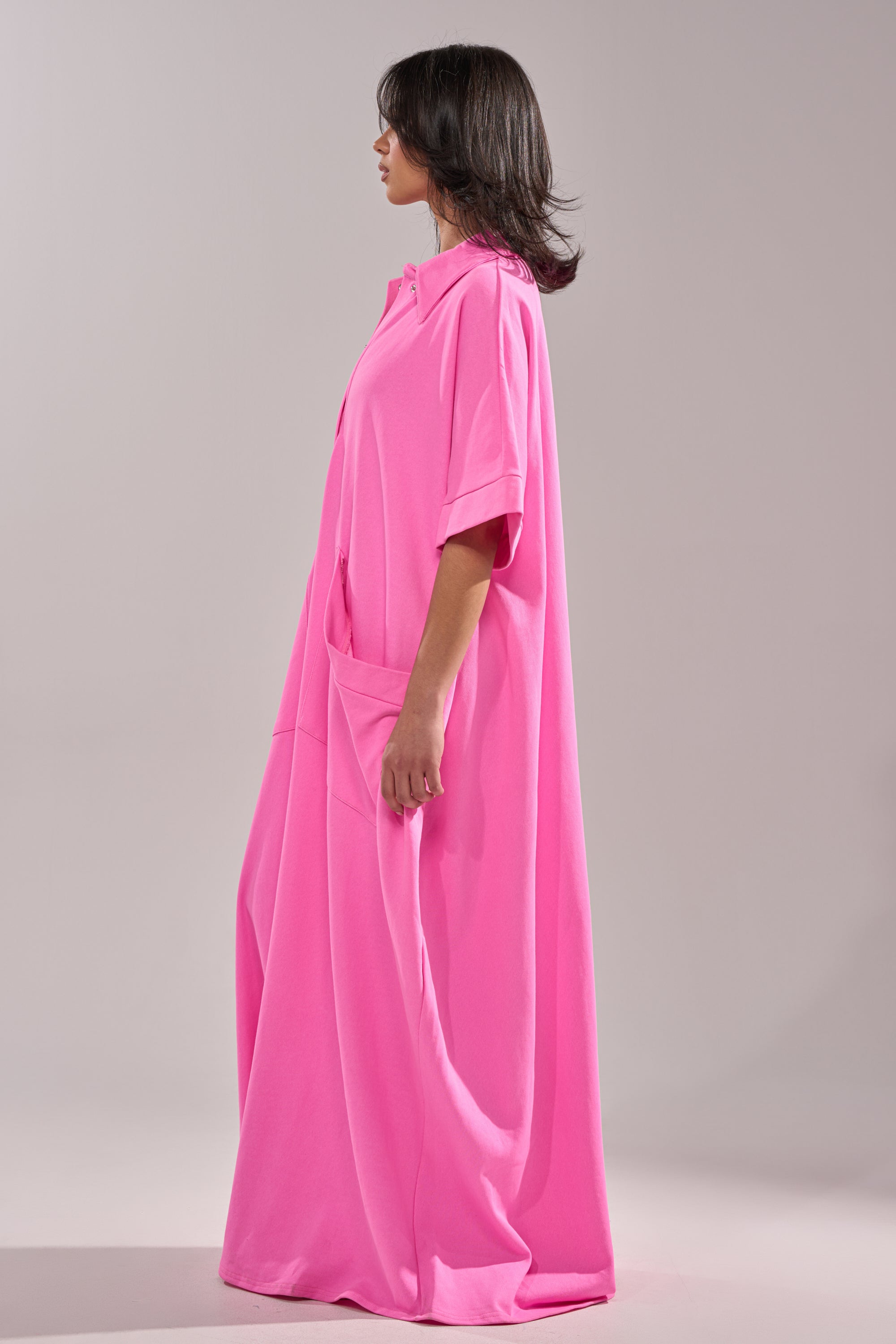 A woman stands in profile against a plain light background wearing the PRETTY IN PINK JUMPSUIT—an oversized, bright pink, floor-length one-piece with short sleeves and a collar, ideal for resort wear.