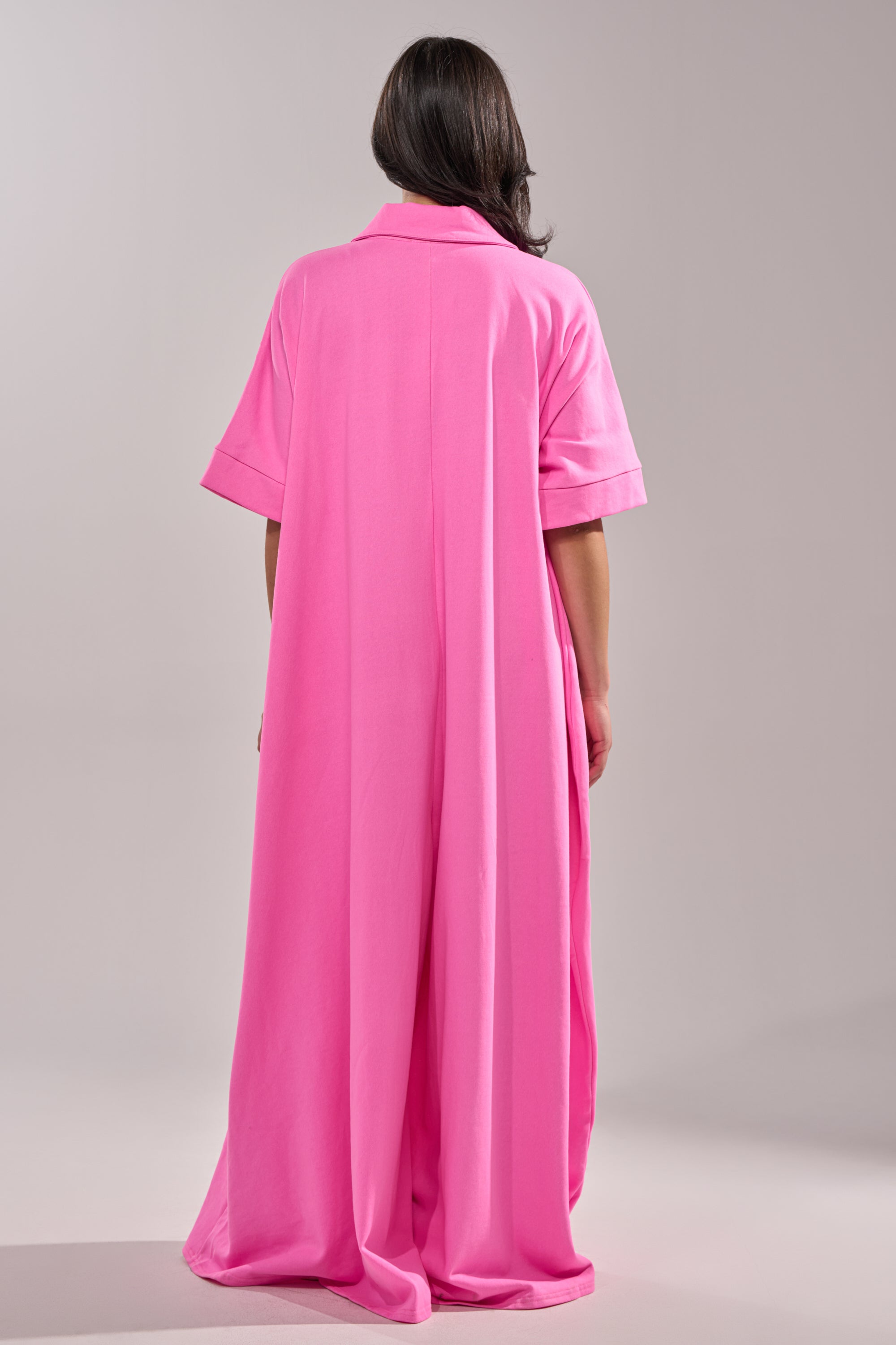 A person faces away, wearing the PRETTY IN PINK JUMPSUIT—a loose, floor-length, bright pink one-piece with short sleeves and a collar—against a plain, light-colored background.