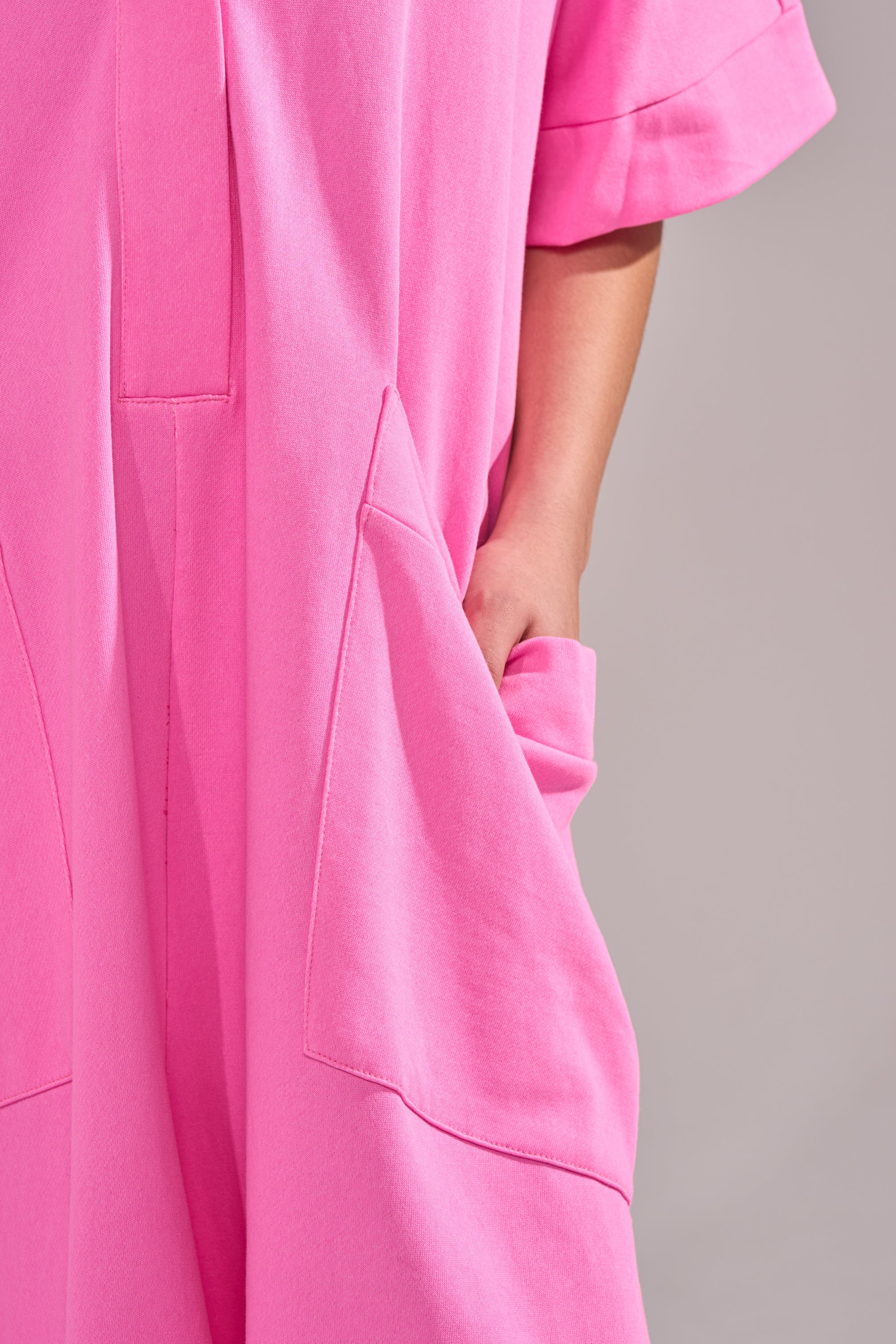 A person models the PRETTY IN PINK JUMPSUIT, featuring a bright pink hue, wide-leg silhouette, and large pockets, standing with one hand in a pocket against a neutral background.