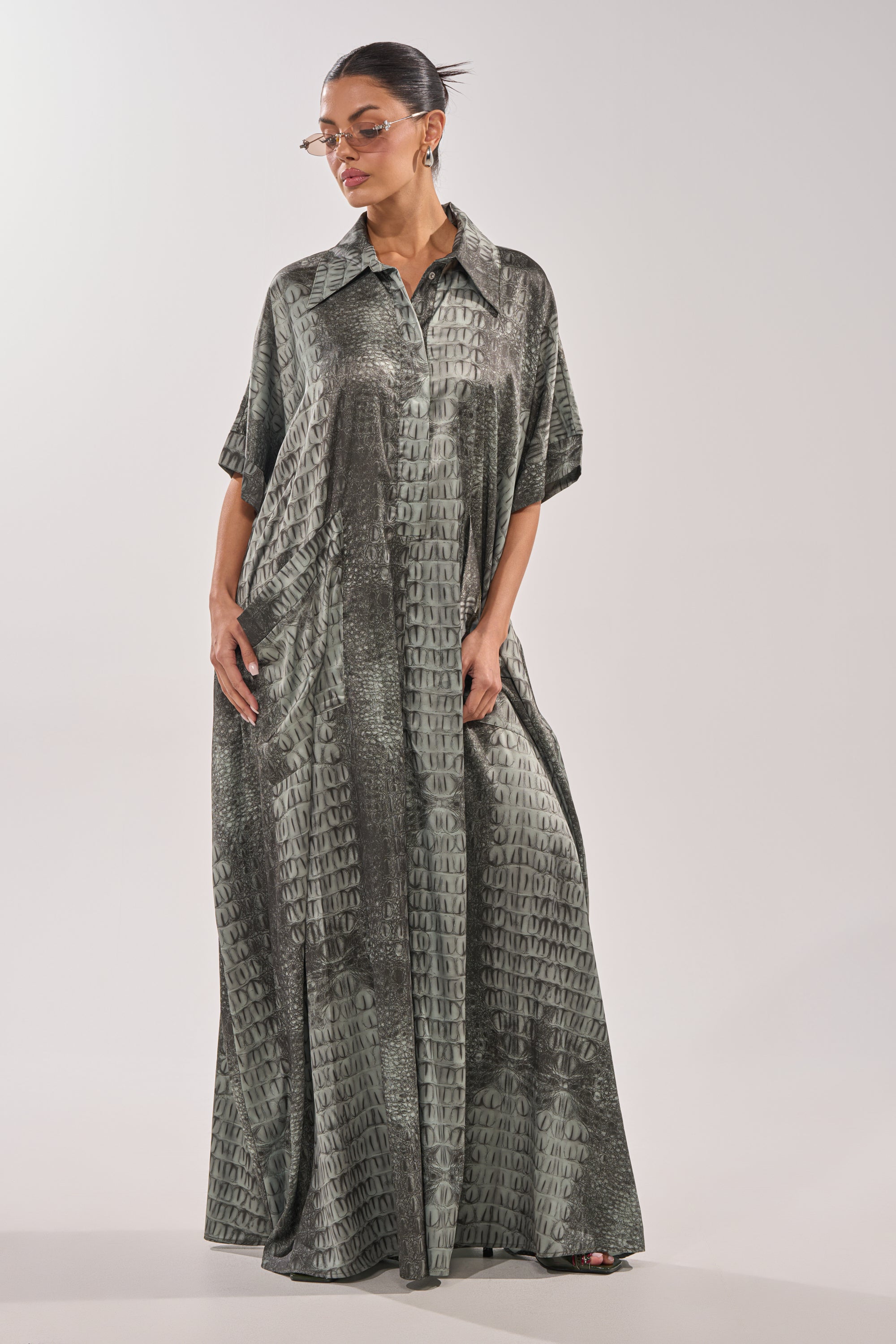 A woman stands against a plain background, looking to the side with her hands in the pockets of the SHE'S RICH OVERSIZED JUMPSUIT IN SNAKE, styled with a collar, short sleeves, and sunglasses. 0