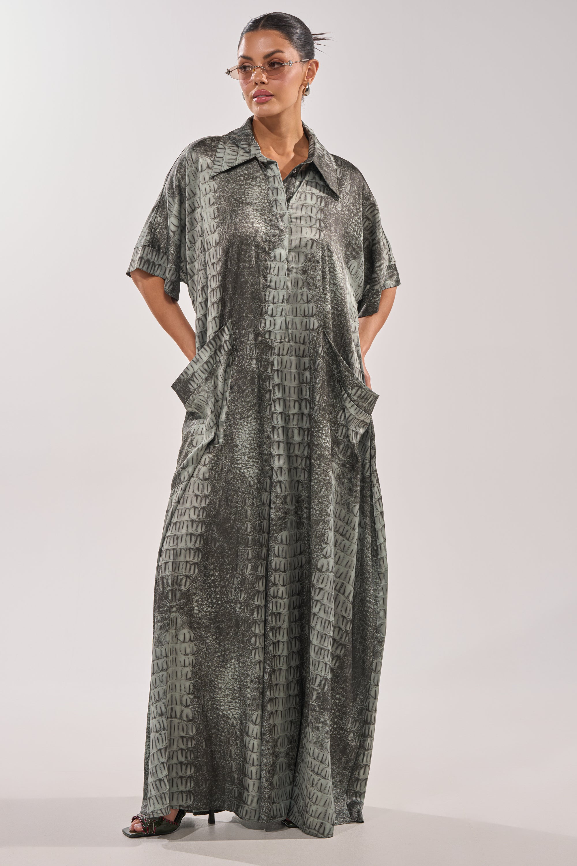 A woman wears the SHE'S RICH OVERSIZED JUMPSUIT IN SNAKE, a wide-leg, ankle-length piece with a textured gray-green pattern. She stands hands-in-pockets, hair tied back, accessorized with sunglasses and heeled sandals. 1