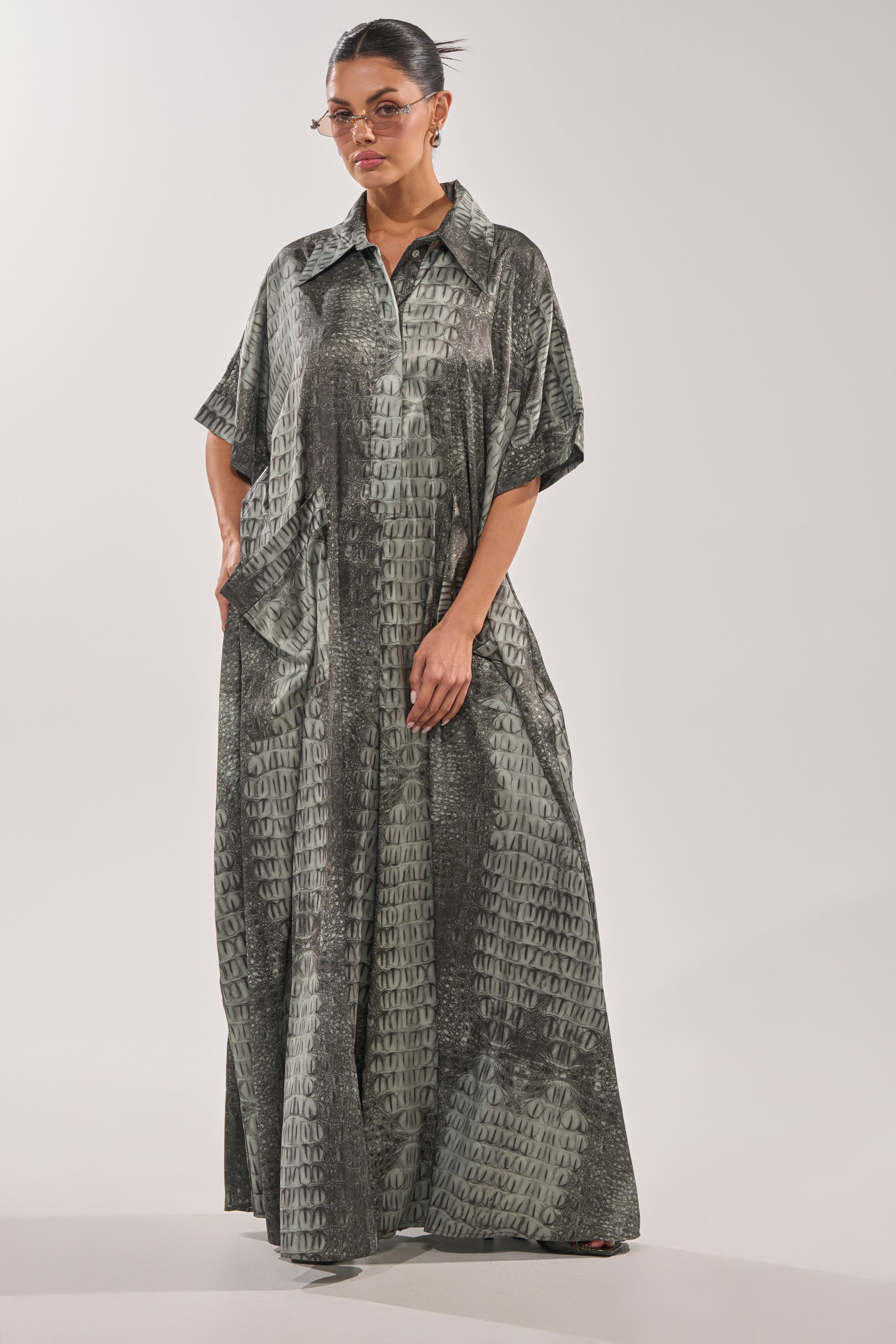A woman in the SHE'S RICH OVERSIZED JUMPSUIT IN SNAKE, featuring a green-gray snakeskin print, collar, short sleeves, pockets, and a loose floor-length fit, stands against a plain white background while wearing sunglasses. 2