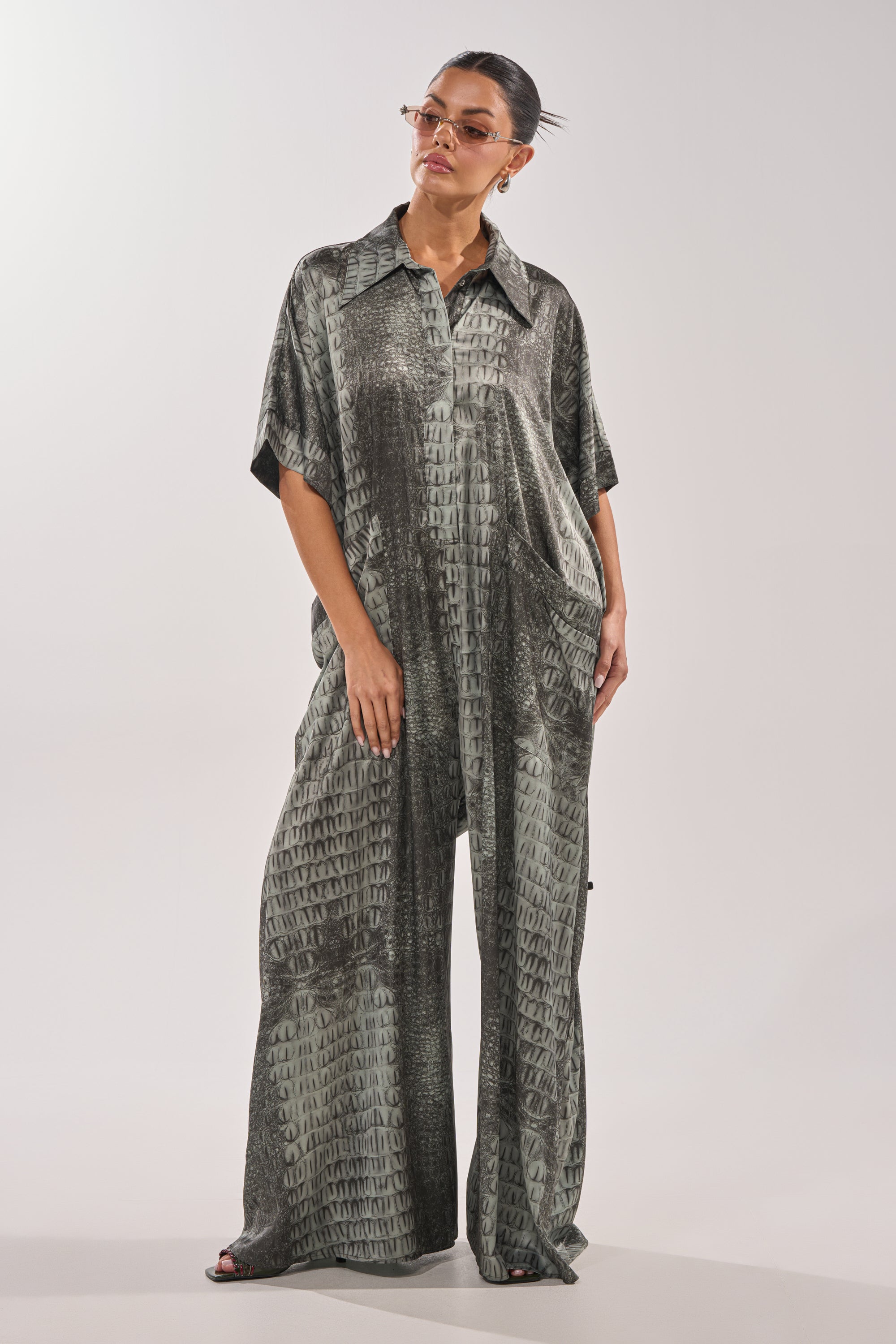 A woman in the SHE'S RICH OVERSIZED JUMPSUIT IN SNAKE, wearing oversized sunglasses and standing with her hands in her pockets, gazes to the side against a plain background. 3