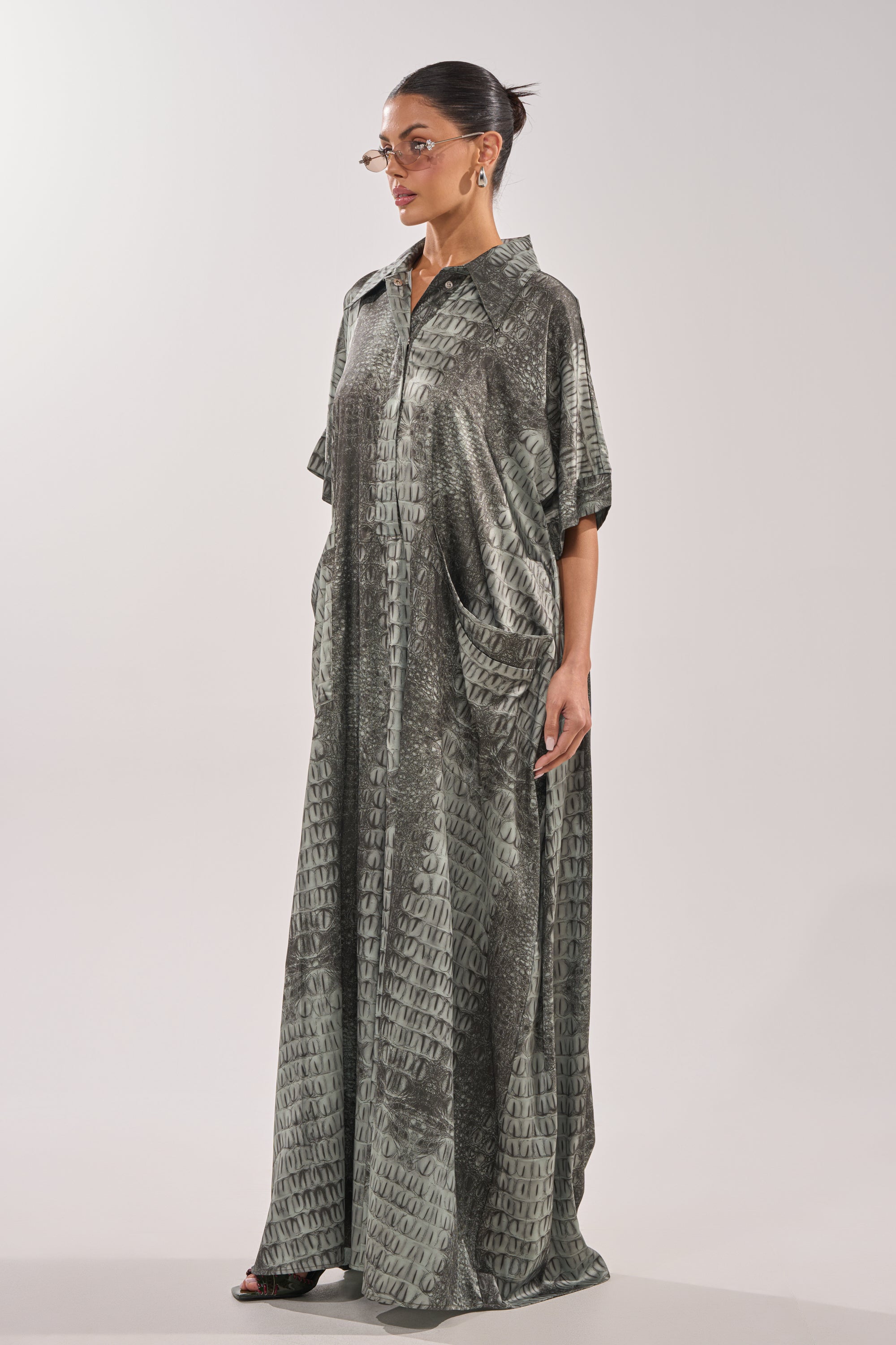 A person wearing the SHE'S RICH OVERSIZED JUMPSUIT IN SNAKE, a loose-fitting, gray patterned design with short sleeves and front pockets, stands against a plain light background, accessorized with glasses and earrings. 4