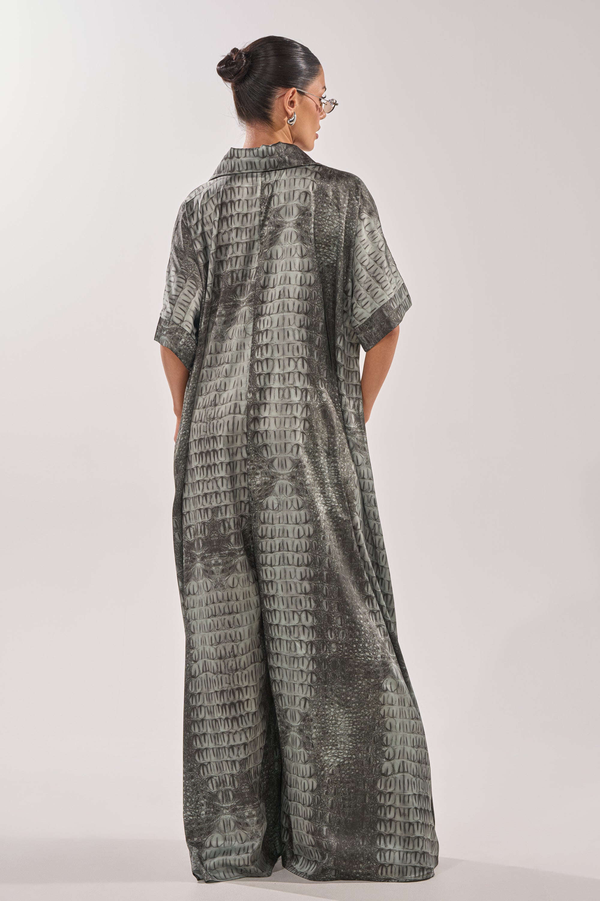A woman with dark hair in a bun and glasses models the SHE'S RICH OVERSIZED JUMPSUIT IN SNAKE, facing away from the camera against a plain light background. 5