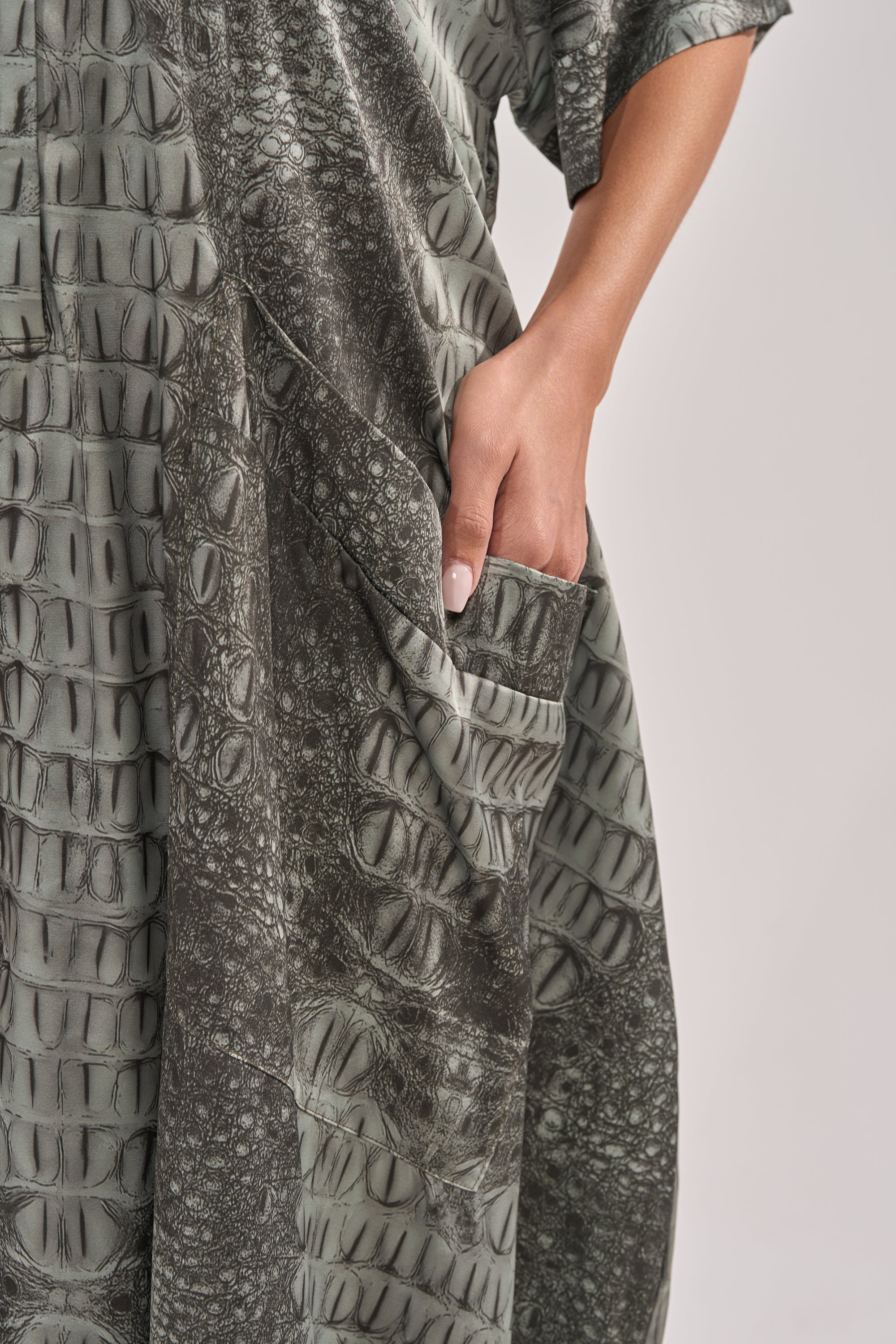 A person wearing the SHE'S RICH OVERSIZED JUMPSUIT IN SNAKE places their right hand in the pocket, showcasing the snakeskin print and textured fabric against a plain light background. 6