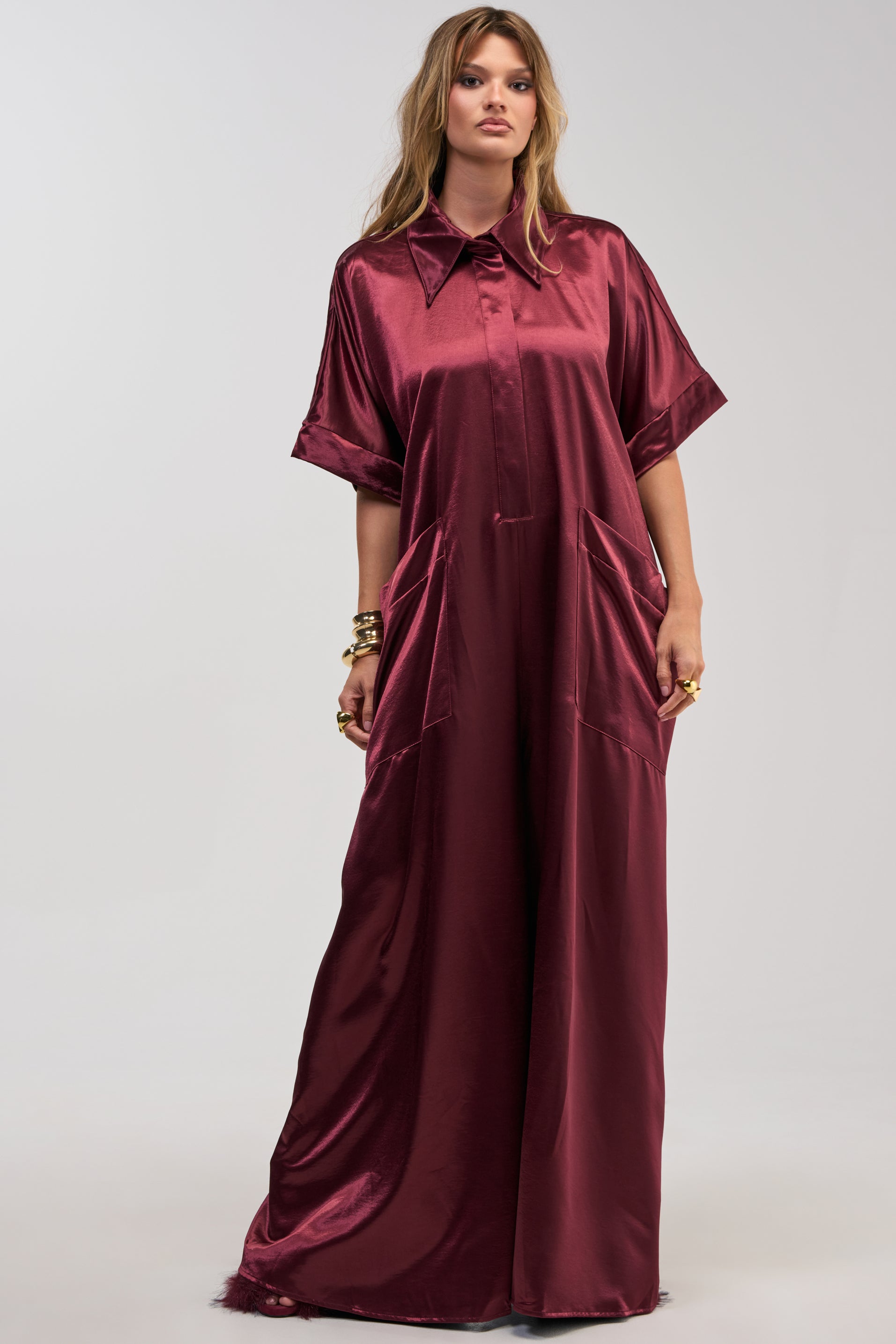 A woman stands against a plain light background wearing the SHE’S RICH OVERSIZED JUMPSUIT IN BURGUNDY, a loose, floor-length satin jumpsuit with short sleeves, a collar, and large pockets.