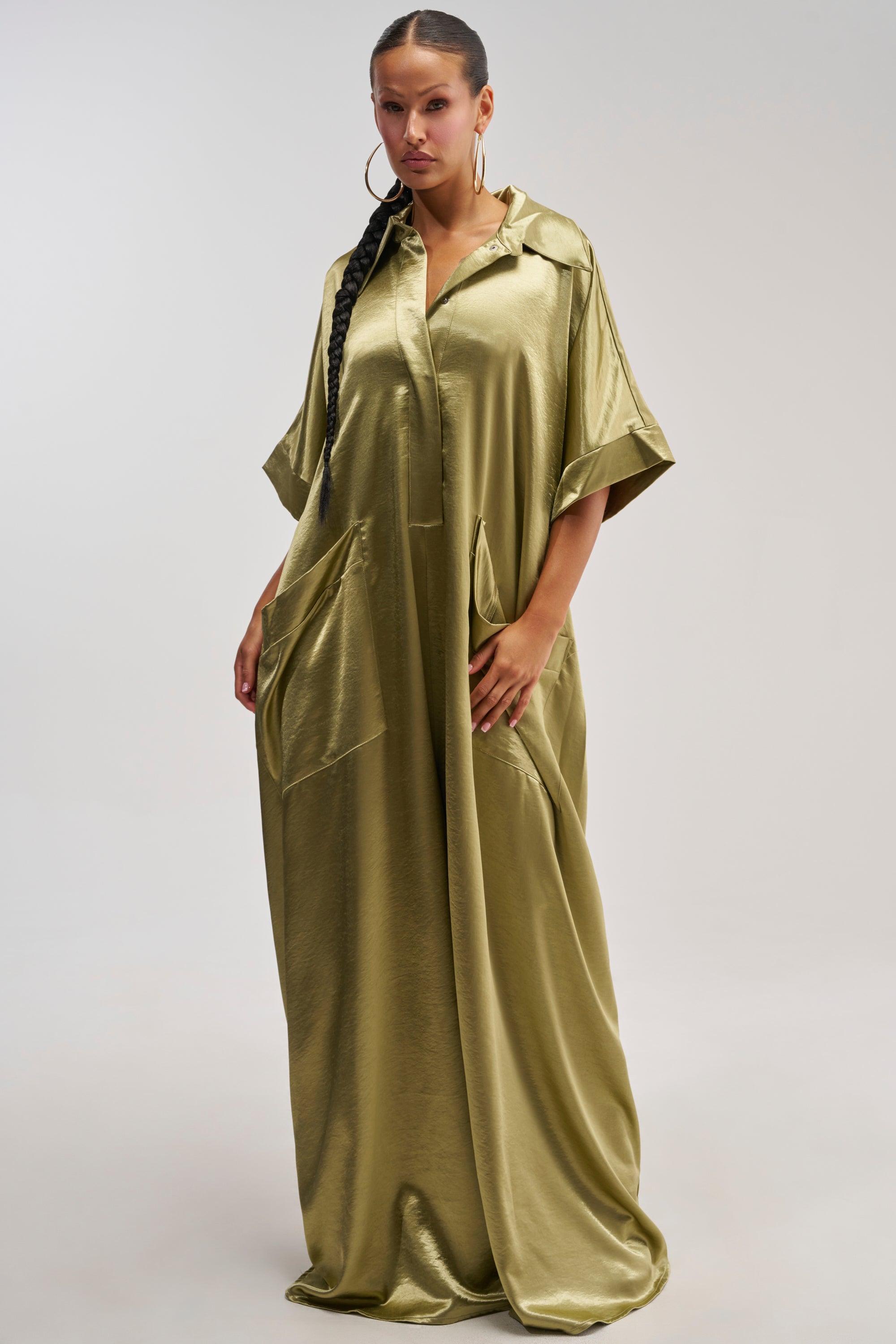 A woman stands against a plain light background, effortlessly capturing the essence of summer styling in the SHE'S RICH OVERSIZED JUMPSUIT IN OLIVE, featuring a loose fit, short sleeves, and large pockets.