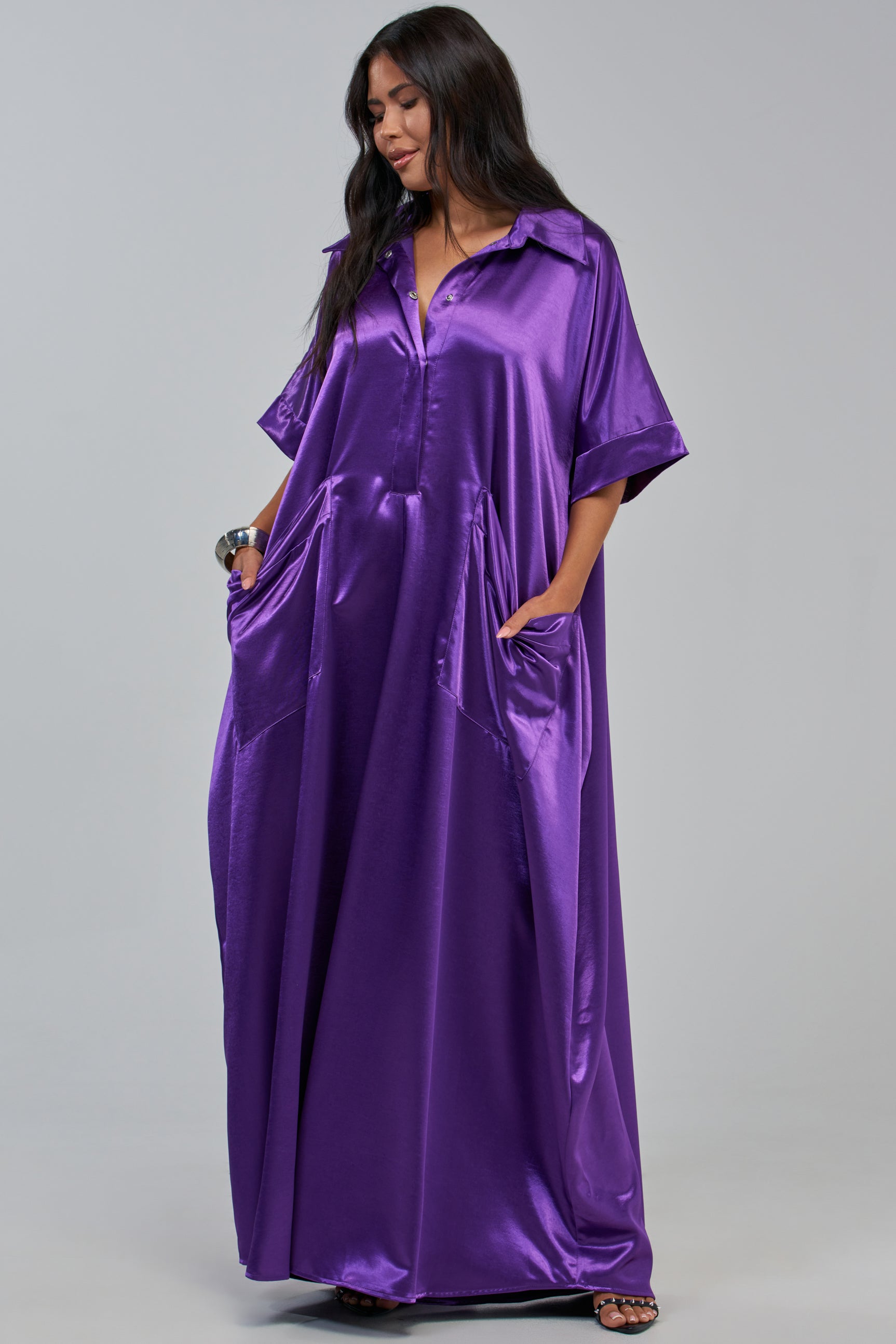 Dressed in the SHE’S RICH OVERSIZED JUMPSUIT IN PURPLE—a loose, shiny, satin piece with short sleeves and pockets—she stands with her hands tucked comfortably in her pockets, looking down and smiling. This look effortlessly blends the elegance of a maxi dress with the comfort and wide-leg style of an oversized jumpsuit.