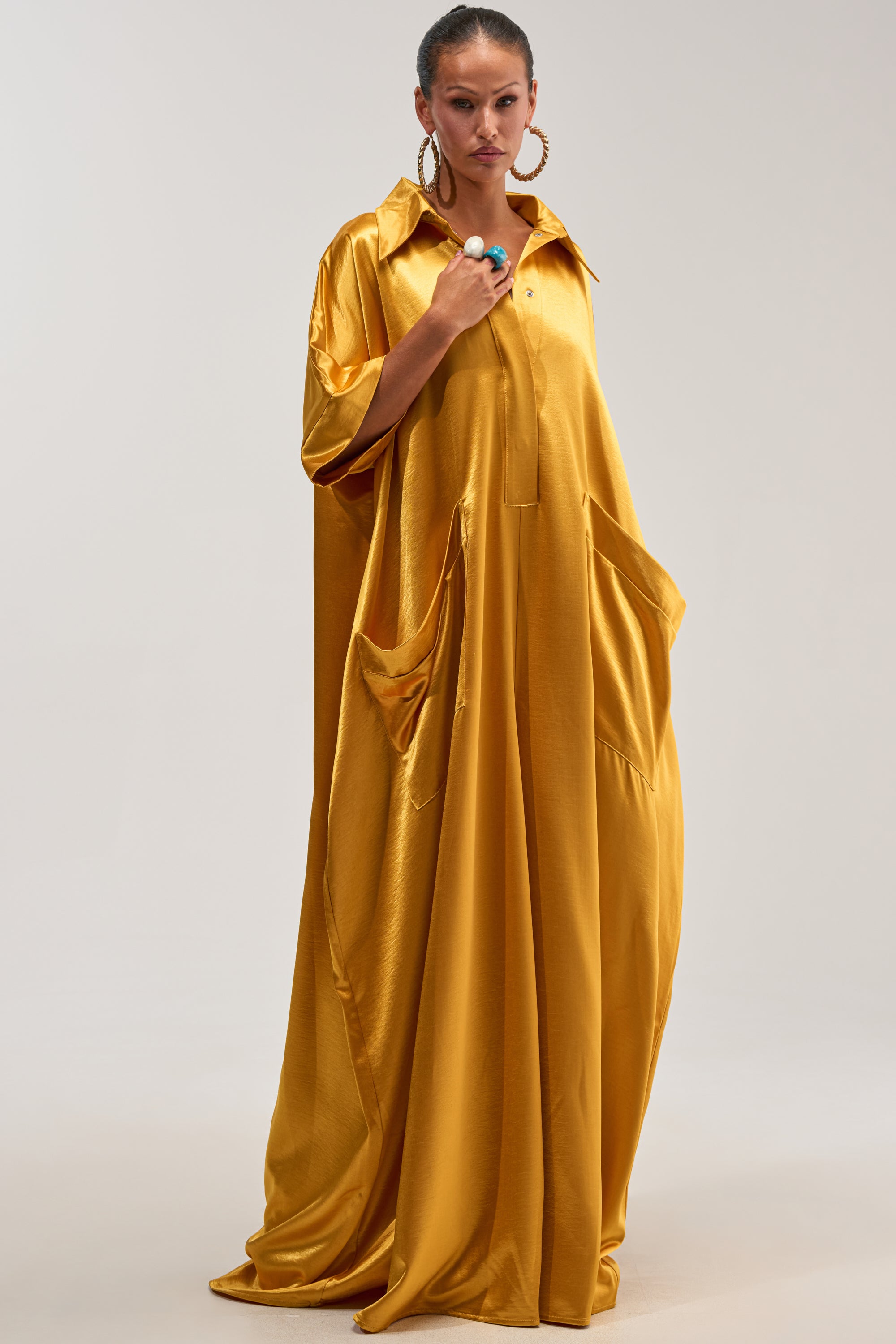 A person wearing the SHE'S RICH OVERSIZED JUMPSUIT IN MUSTARD, featuring a loose fit, collared neckline, large pockets, and rolled sleeves, stands against a plain light background accessorized with hoop earrings and a bold ring.