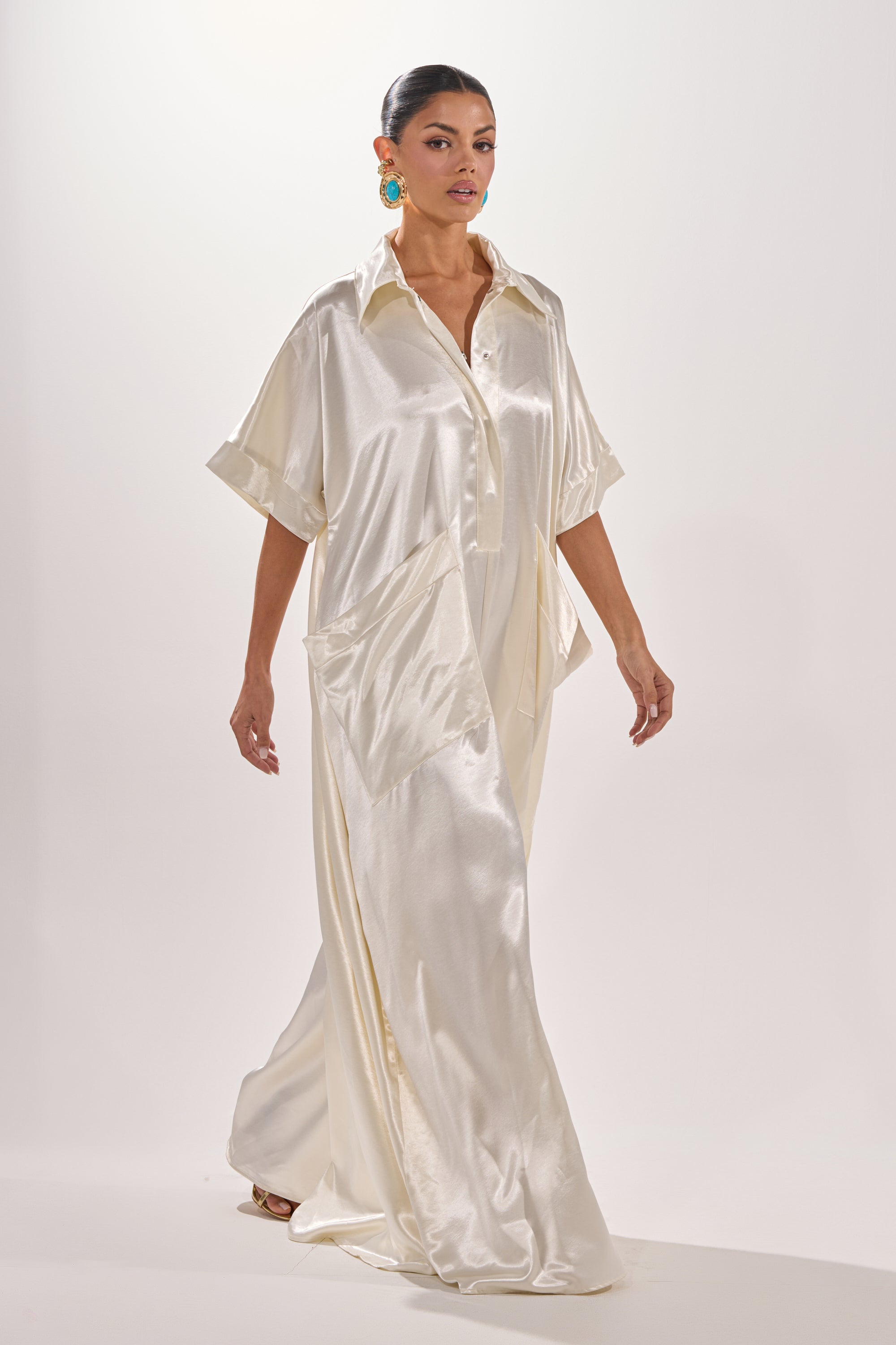 A woman walks confidently in the SHE'S RICH OVERSIZED JUMPSUIT IN LUXE CREAM, featuring a collared neckline and short sleeves, paired with bold turquoise earrings against a plain white background. 0