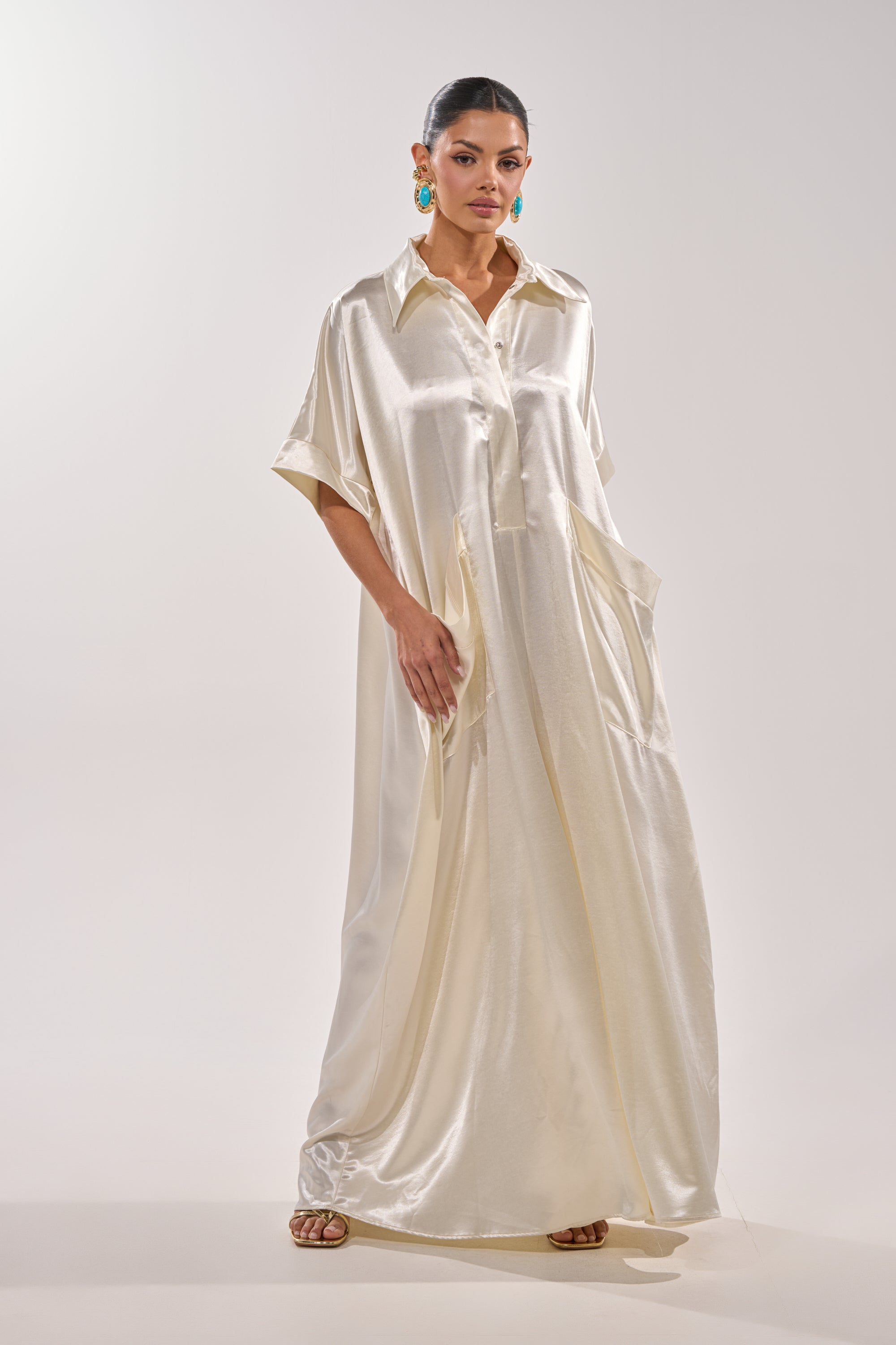 A woman poses against a light background in the SHE'S RICH OVERSIZED JUMPSUIT IN LUXE CREAM—an elegant, loose satin piece with short sleeves and a collar—teamed with turquoise earrings and sandals for a chic minimalist vibe. 1