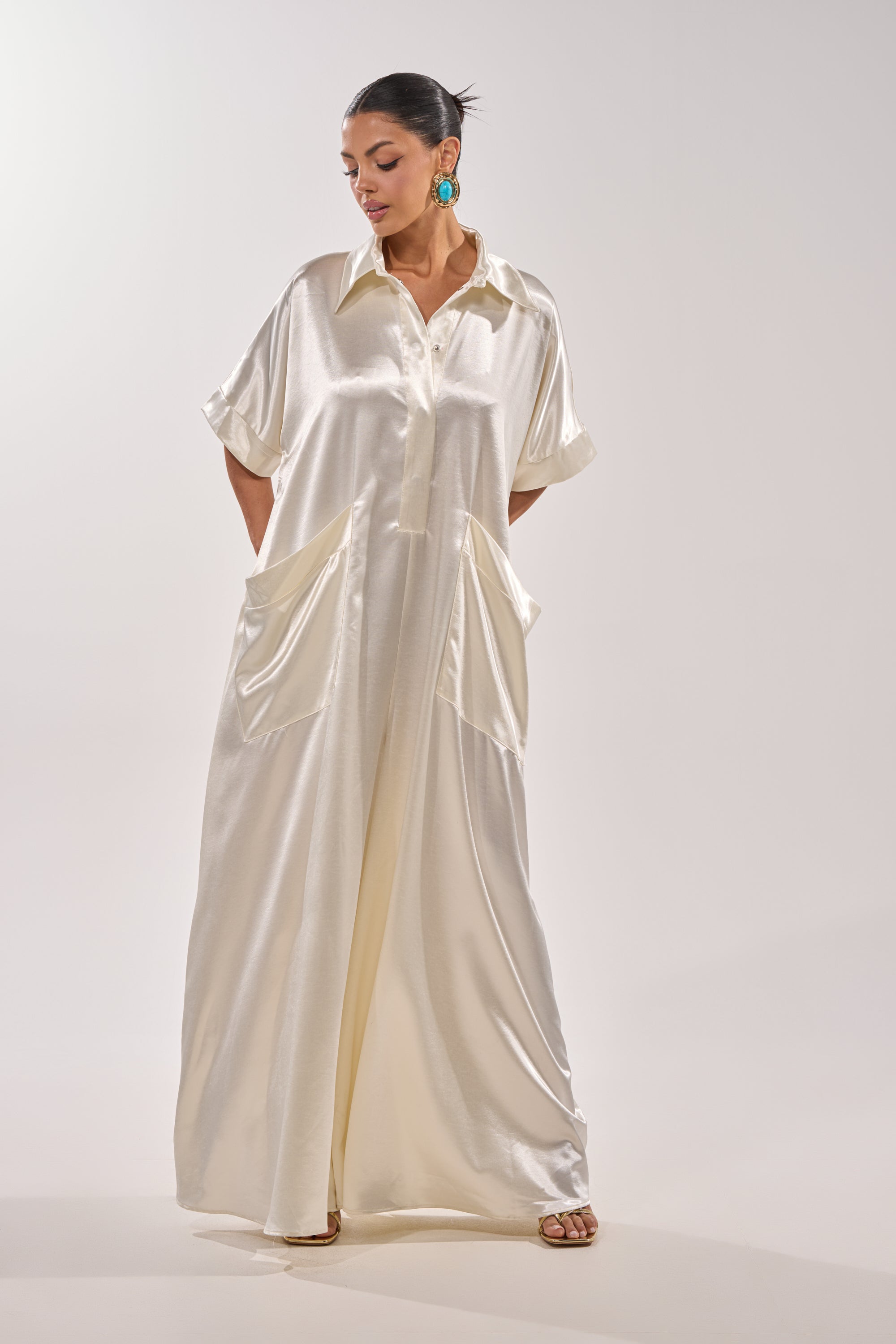 A woman poses in the SHE'S RICH OVERSIZED JUMPSUIT IN LUXE CREAM, featuring a collared neckline, short sleeves, large front pockets, and wide legs. She accessorizes with green statement earrings and has her hair pulled back. 2