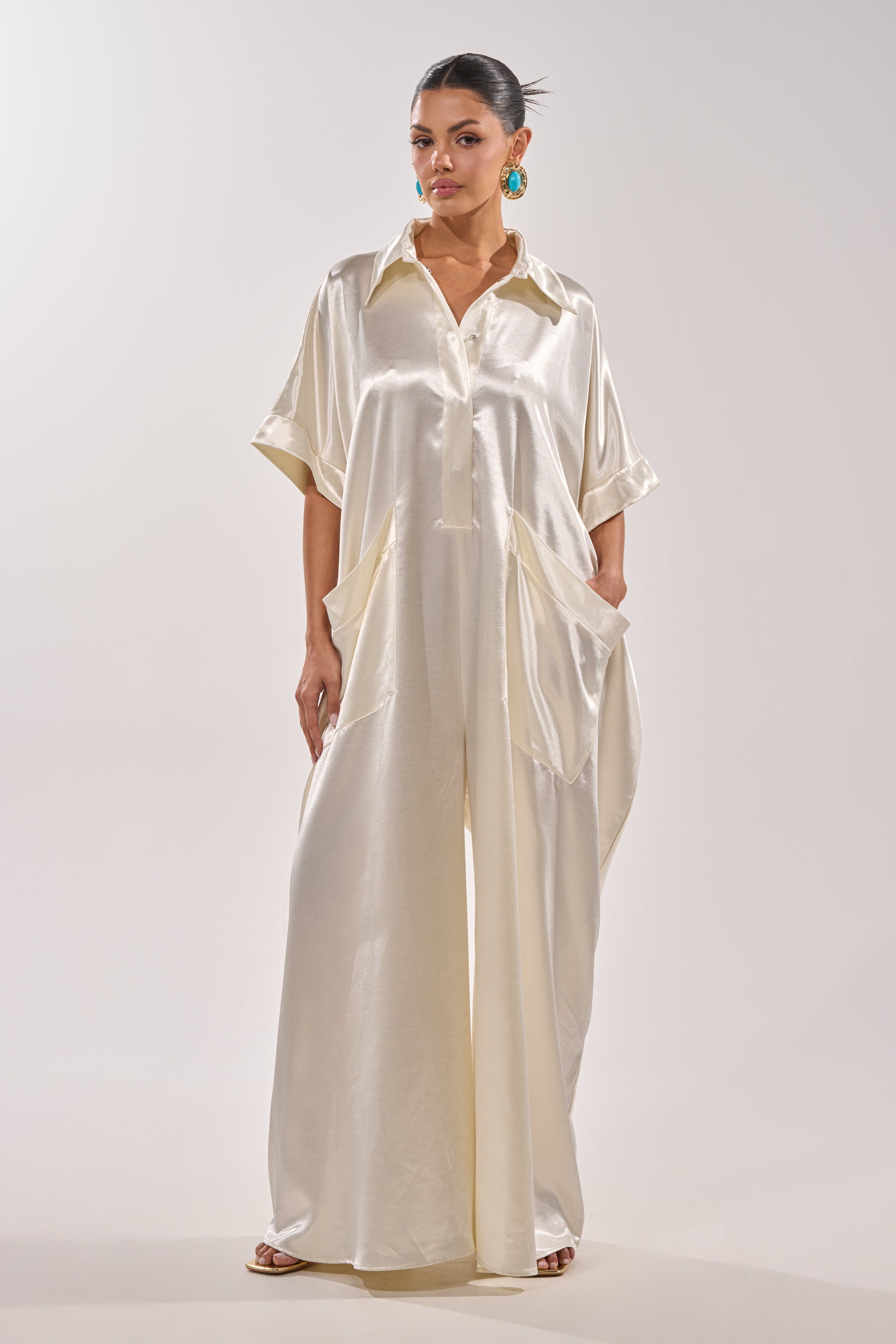 A woman wears the SHE'S RICH OVERSIZED JUMPSUIT IN LUXE CREAM, featuring short sleeves, a collar, wide legs, and large front pockets. She pairs it with blue earrings and open-toe sandals against a plain background. 3