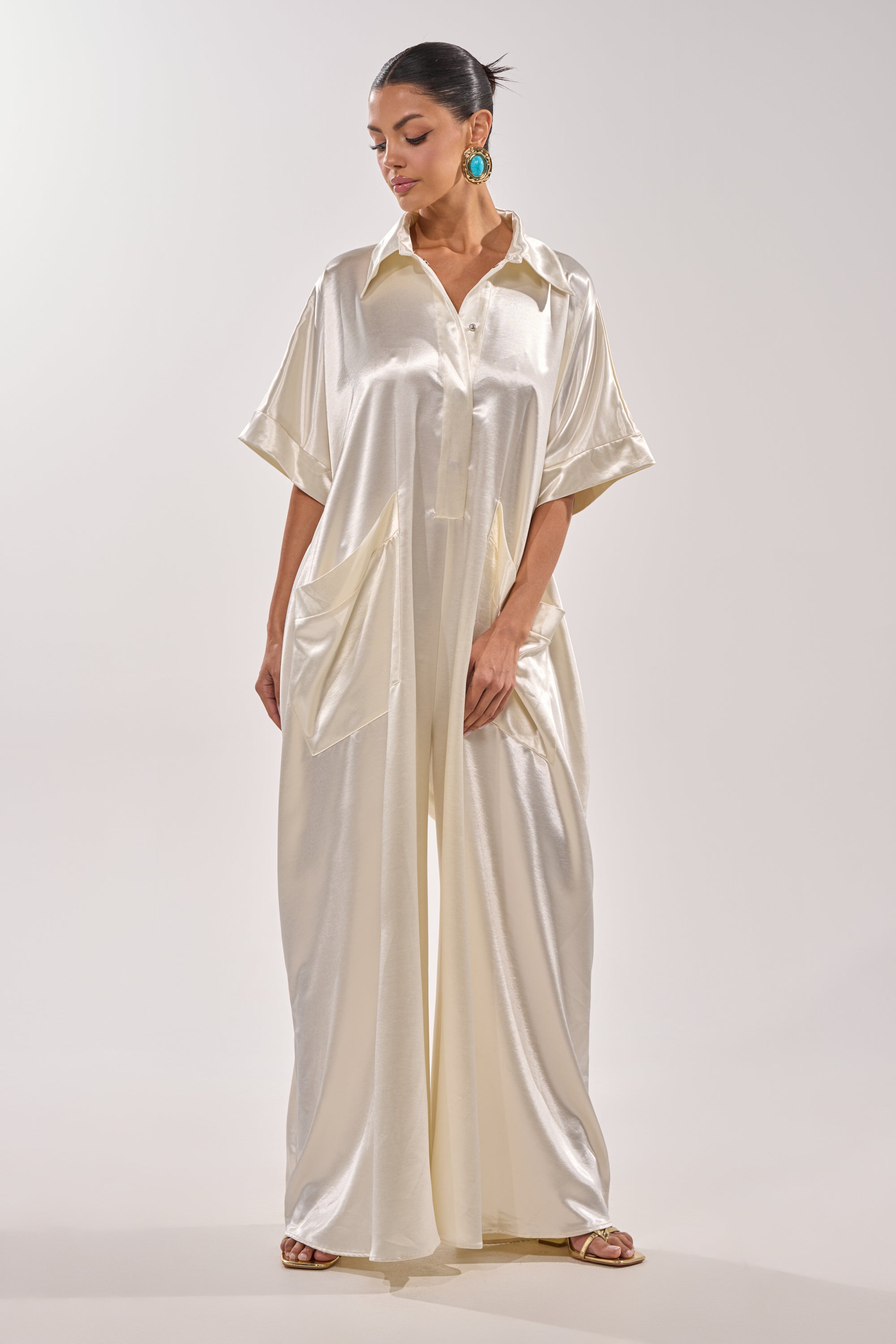 Wearing the SHE'S RICH OVERSIZED JUMPSUIT IN LUXE CREAM with wide legs, short sleeves, and front pockets, a woman styles her pulled-back hair with turquoise statement earrings and brown sandals. 4