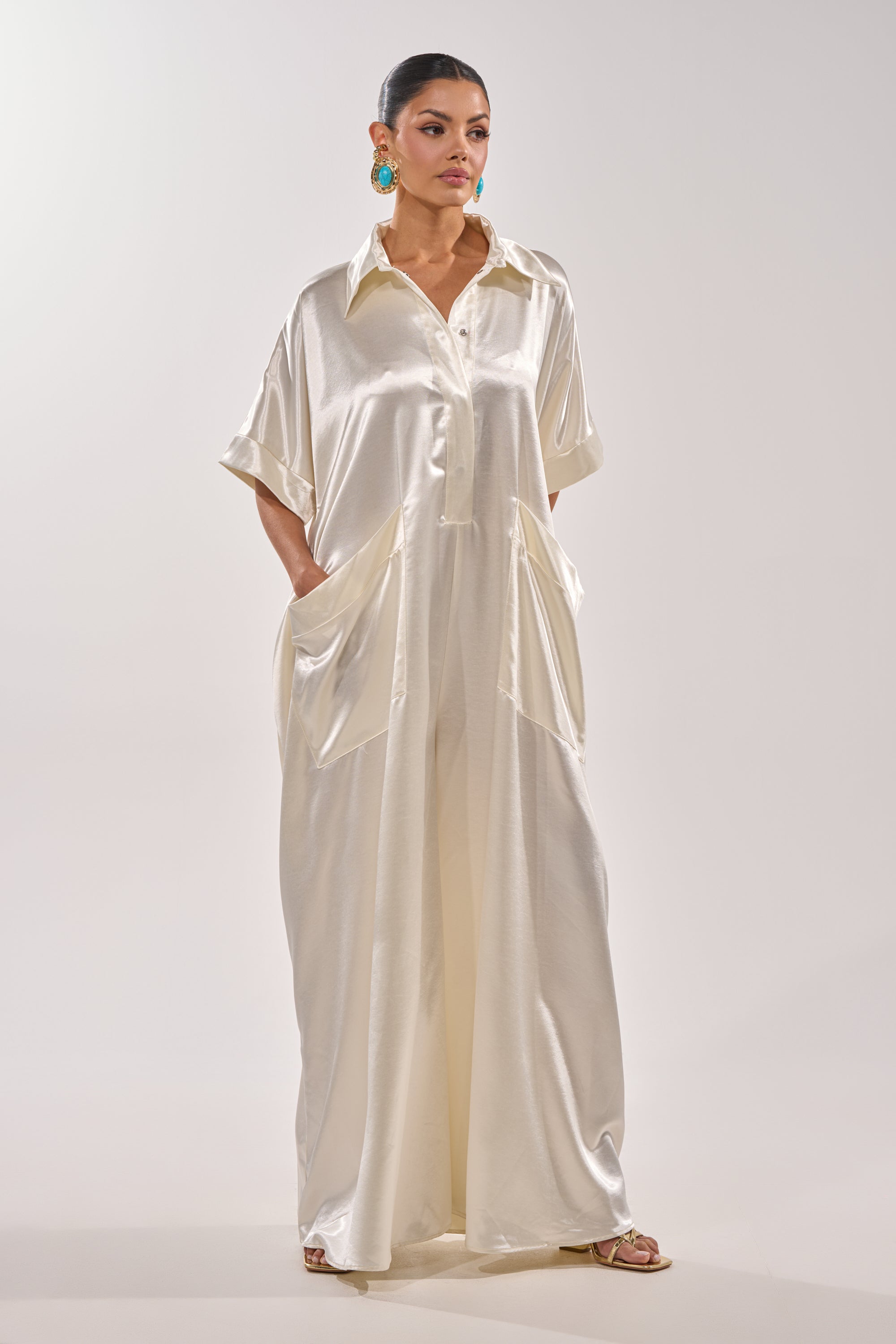 A woman wears the SHE'S RICH OVERSIZED JUMPSUIT IN LUXE CREAM, featuring a loose fit, collared neckline, short sleeves, large pockets, and a maxi silhouette. She styles it with statement blue earrings and open-toed sandals; her hair is tied back. 5