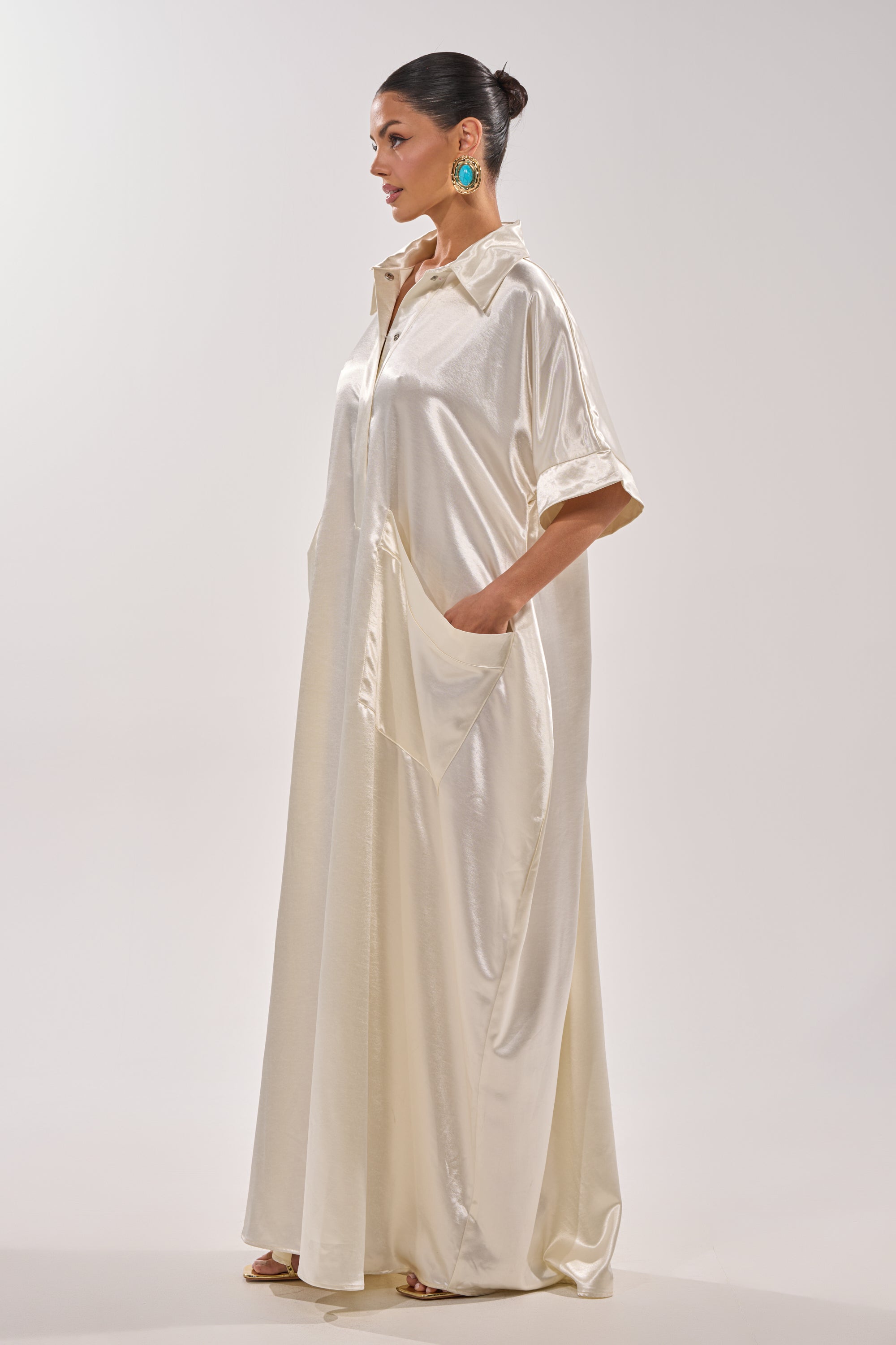 A woman in profile wears the SHE'S RICH OVERSIZED JUMPSUIT IN LUXE CREAM, a long, flowing satin piece with short sleeves and large side pockets. She styles her hair in a neat bun with turquoise earrings and open-toed heels. 6