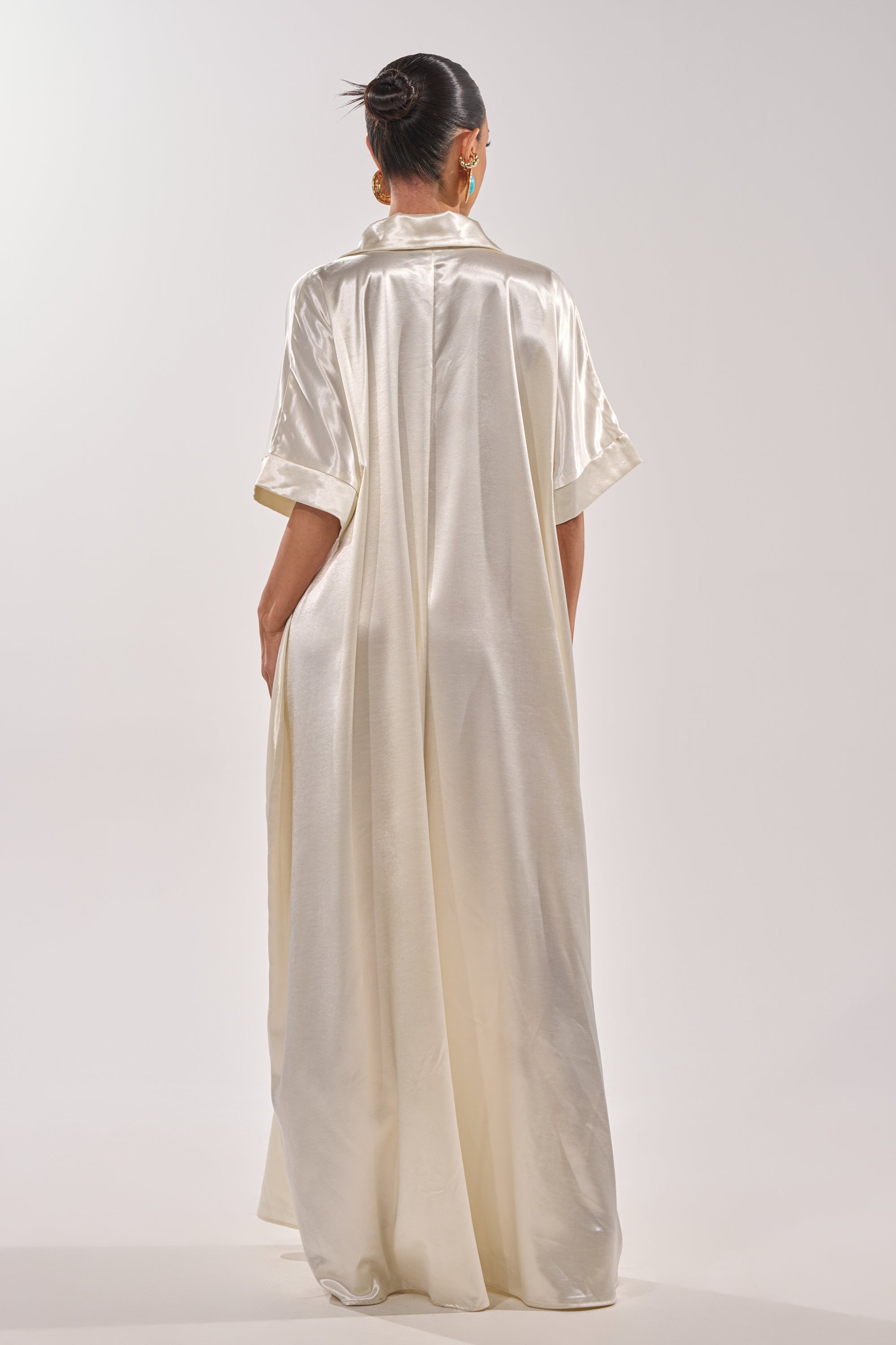 A person stands with their back to the camera in the SHE'S RICH OVERSIZED JUMPSUIT IN LUXE CREAM. The flowing, satin jumpsuit features short sleeves and drapes to the floor. Their hair is in a sleek bun against a plain, light background. 7