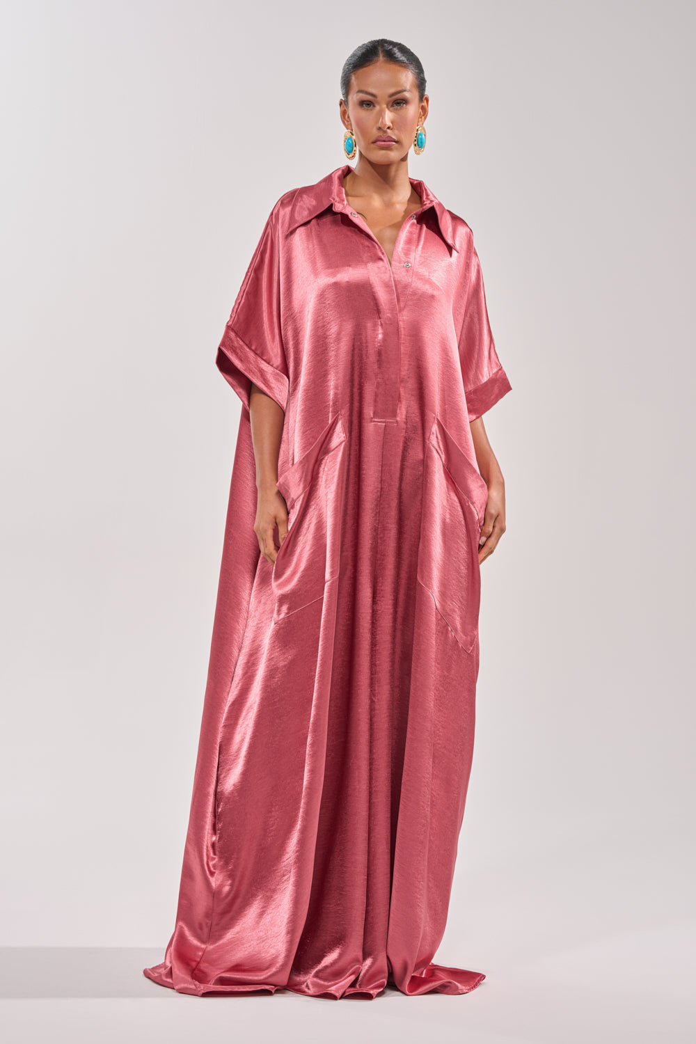 A woman wears the SHE'S RICH OVERSIZED JUMPSUIT IN ROSE—a loose, floor-length satin jumpsuit with short sleeves, a collar, and large pockets—styling it with colorful earrings for a modern evening look against a plain backdrop. 1