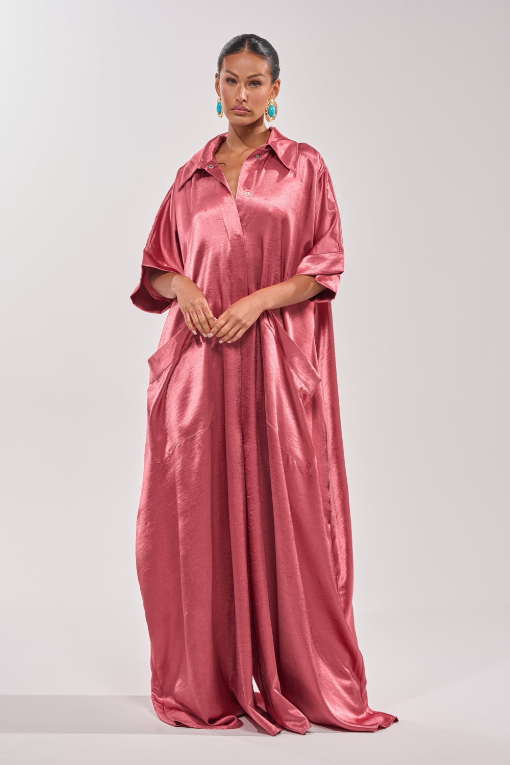 A woman wears the SHE'S RICH OVERSIZED JUMPSUIT IN ROSE, a loose, floor-length satin piece with wide sleeves and large pockets. She pairs it with colorful earrings, standing against a light gray background. 2