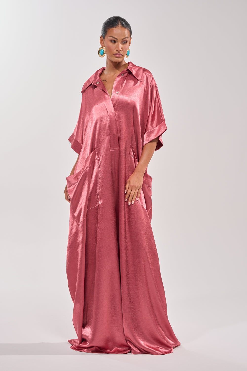 A woman poses confidently against a plain light background, wearing the SHE'S RICH OVERSIZED JUMPSUIT IN ROSE—an airy, short-sleeved satin jumpsuit with large pockets. Her hair is pulled back and she accessorizes with bold turquoise earrings. 3