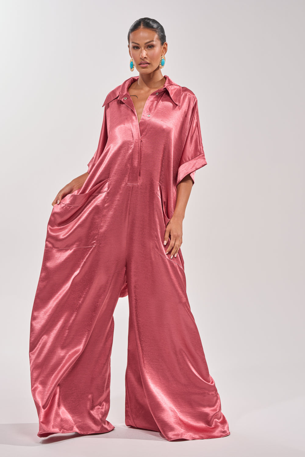 A woman poses in the SHE'S RICH OVERSIZED JUMPSUIT IN ROSE, a loose-fitting satin wide-leg style with short sleeves, collar, and roomy side pockets. She pairs it with turquoise earrings against a light background—perfect for chic evenings. 4