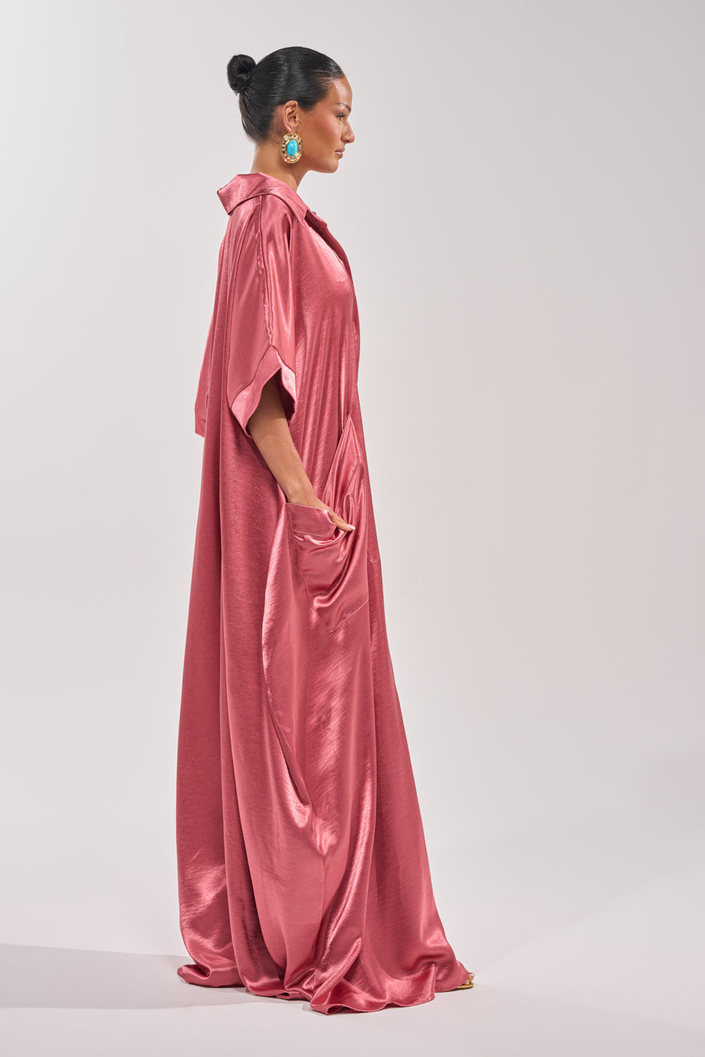 A woman stands in profile wearing the SHE'S RICH OVERSIZED JUMPSUIT IN ROSE, a satin pink jumpsuit with short sleeves and pockets. Her hair is in a bun with large turquoise earrings. The background is plain and light-colored. 5