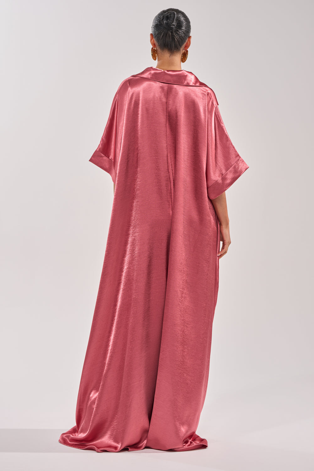 A person stands facing away, wearing the SHE'S RICH OVERSIZED JUMPSUIT IN ROSE with short sleeves. Their hair is in a sleek bun and they wear large round earrings, set against a plain, light-colored background. 6