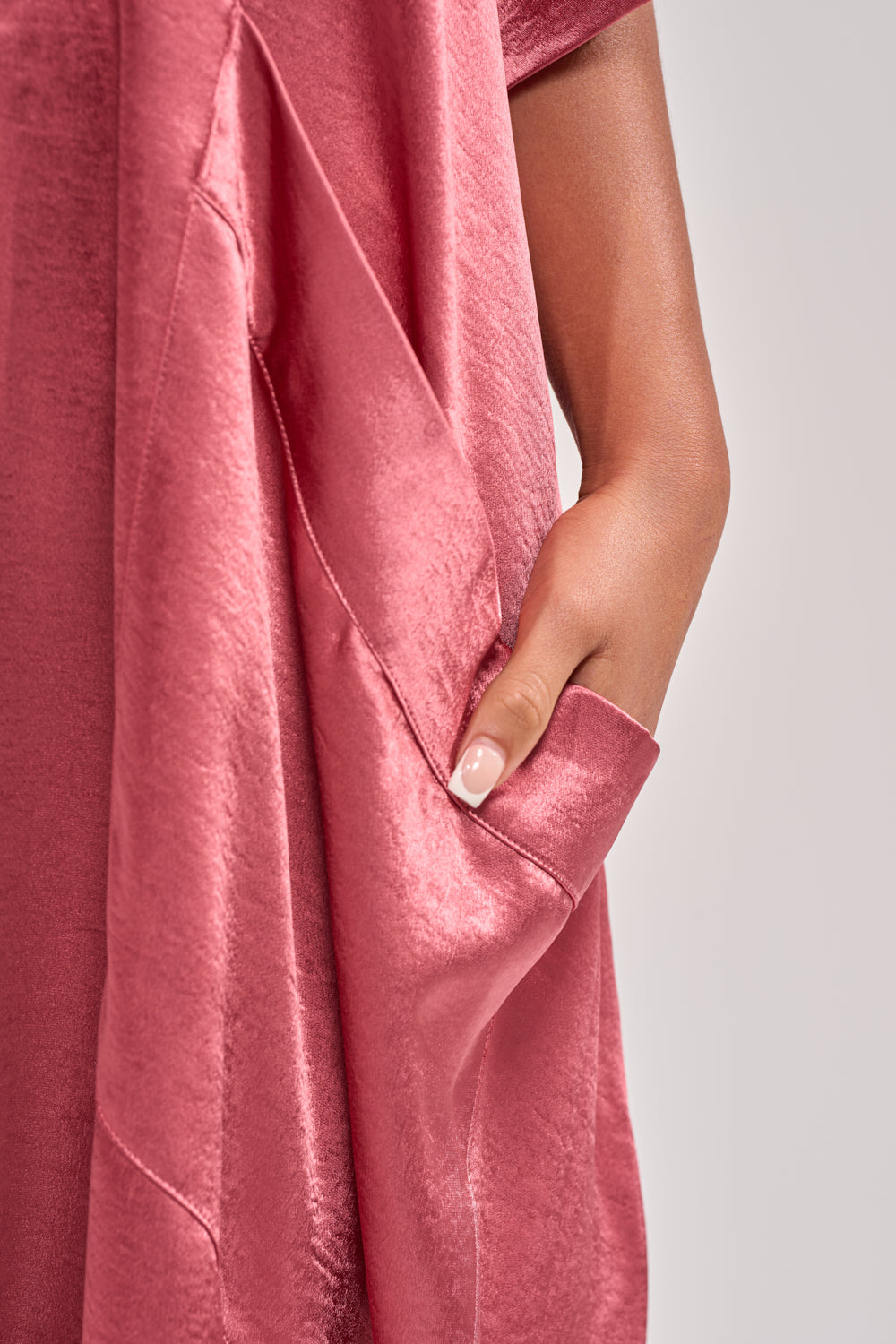 A hand with a light pink manicure slips into the pocket of the SHE'S RICH OVERSIZED JUMPSUIT IN ROSE, its soft, shiny satin fabric standing out against a neutral light gray background for an elegant eveningwear vibe. 7