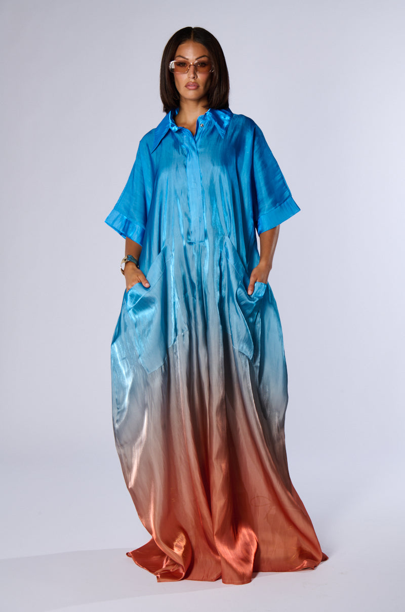 A person stands confidently in the ISLA OMBRE OVERSIZED SATIN JUMPSUIT from AKIRA, which features a striking transition from blue at the collar and sleeves to orange at the hem. They accessorize with sunglasses and have their hands in the jumpsuit's wide pockets, completing their chic summer look against a plain, light-colored background.