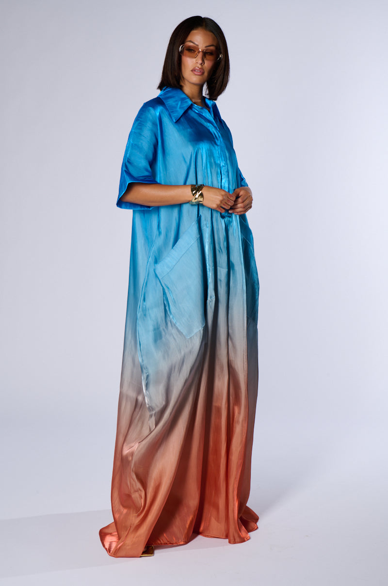 A person stands wearing the ISLA OMBRE OVERSIZED SATIN JUMPSUIT from AKIRA, showcasing a gradient design that transitions from blue at the top to pink and then to an orange hue at the bottom. They have sunglasses on, and their hands are resting on top of a visible pair of pockets, completing the perfect summer look.