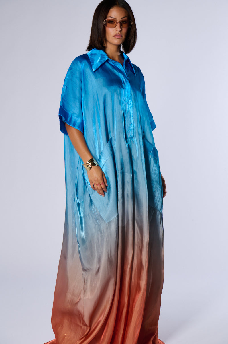 A person stands wearing the ISLA OMBRE OVERSIZED SATIN JUMPSUIT by AKIRA, featuring a gradient pattern that transitions from bright blue at the top to deep red near the bottom. They have long hair and are wearing sunglasses and a bracelet, with hands resting in the jumpsuit pockets—a perfect summer look.