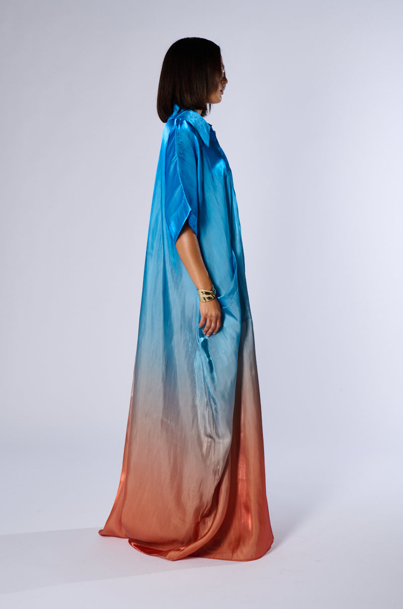 A person with shoulder-length hair faces sideways, wearing the ISLA OMBRE OVERSIZED SATIN JUMPSUIT by AKIRA. The jumpsuit, perfect for a summer look, transitions from blue at the top to orange at the bottom and features short sleeves and a high collar. The individual is posing against a plain white background.