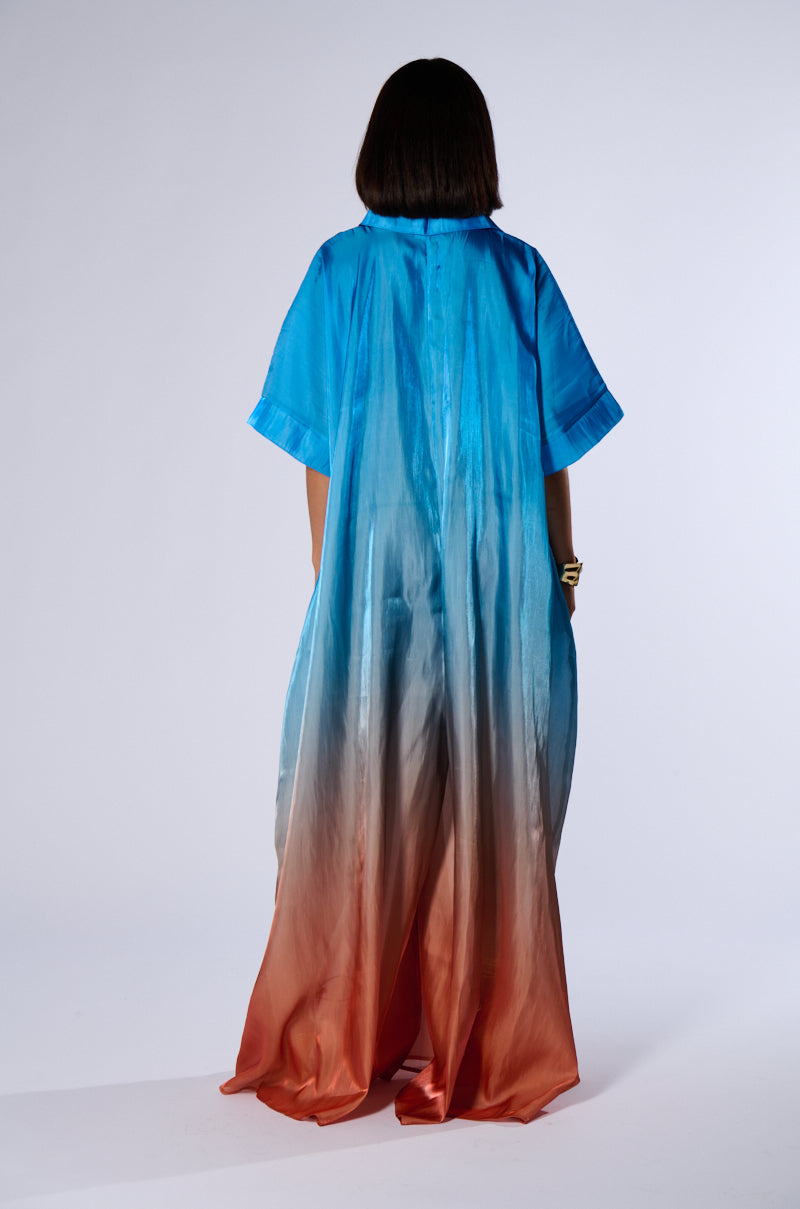 A person with shoulder-length dark hair stands facing away, donning the AKIRA ISLA OMBRE OVERSIZED SATIN JUMPSUIT that transitions from blue at the top to orange at the bottom. The short sleeves add a touch of elegance to their summer look against a plain light grey background.