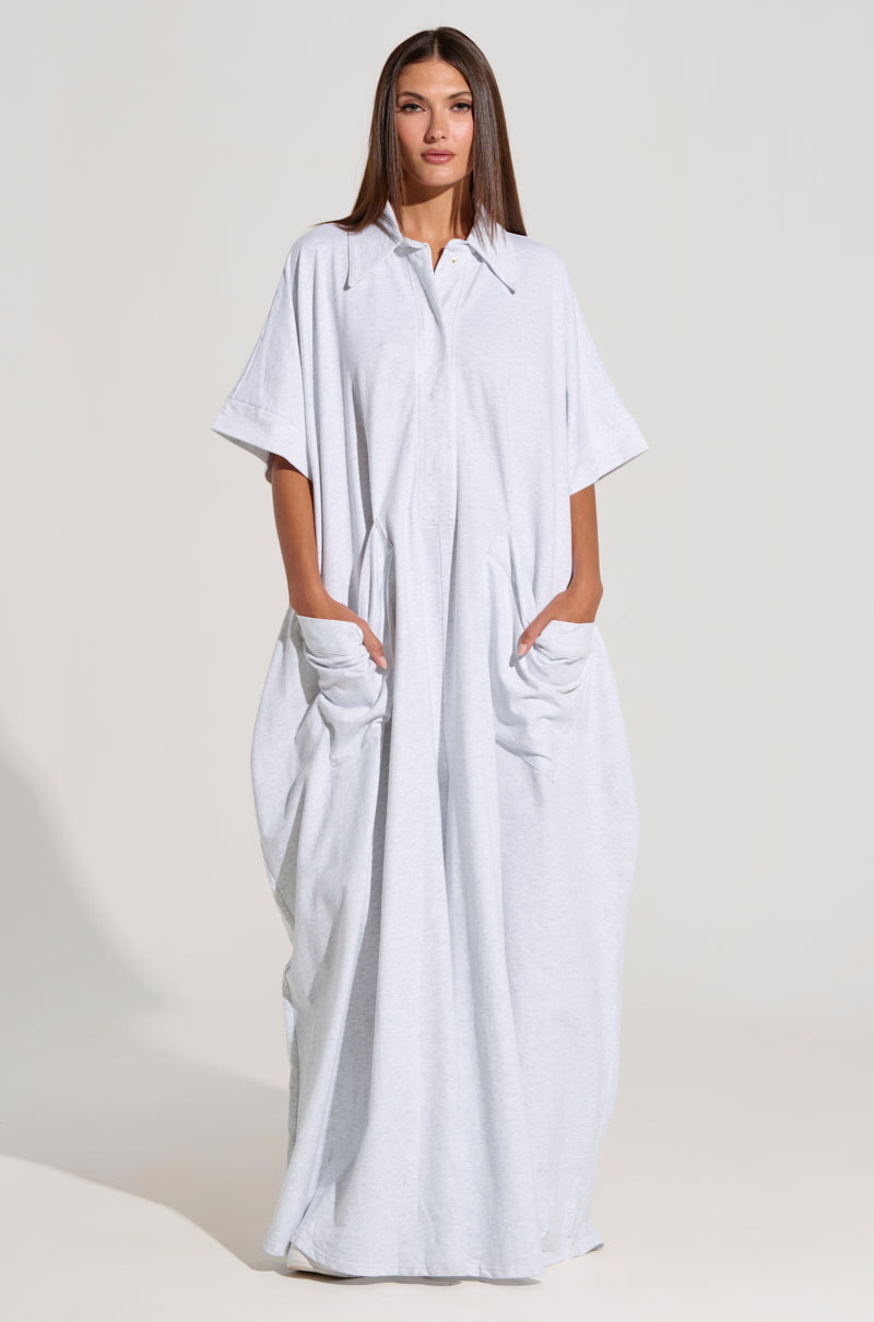 A woman with long, straight hair is wearing the **She’s Rich Oversized Jumpsuit in Heather Grey**, featuring short sleeves and a collar. She has her hands in the jumpsuit's large front pockets. The background is plain and light-colored.