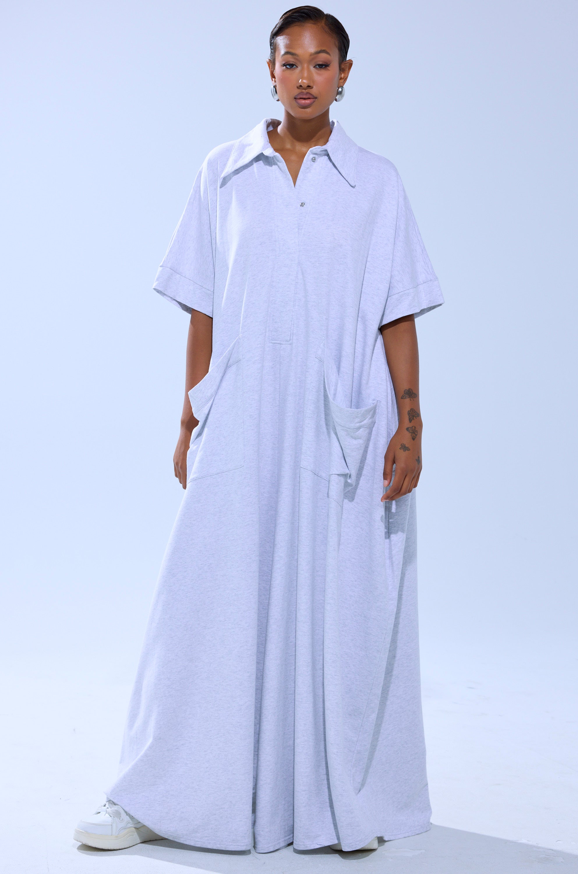 A woman with short hair and visible arm tattoos wears the SHE'S RICH OVERSIZED JUMPSUIT IN HEATHER GREY. The ankle-length, collared piece features short sleeves, large pockets, and pairs with white sneakers against a plain light background.