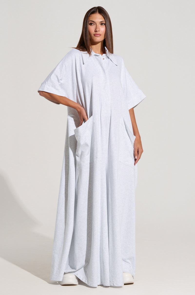 A woman with straight hair stands against a plain background, wearing the SHE'S RICH OVERSIZED JUMPSUIT IN HEATHER GREY. This loose-fitting, full-length piece features short sleeves and large pockets where she rests her hands. She completes her look with white shoes and gazes directly at the camera.