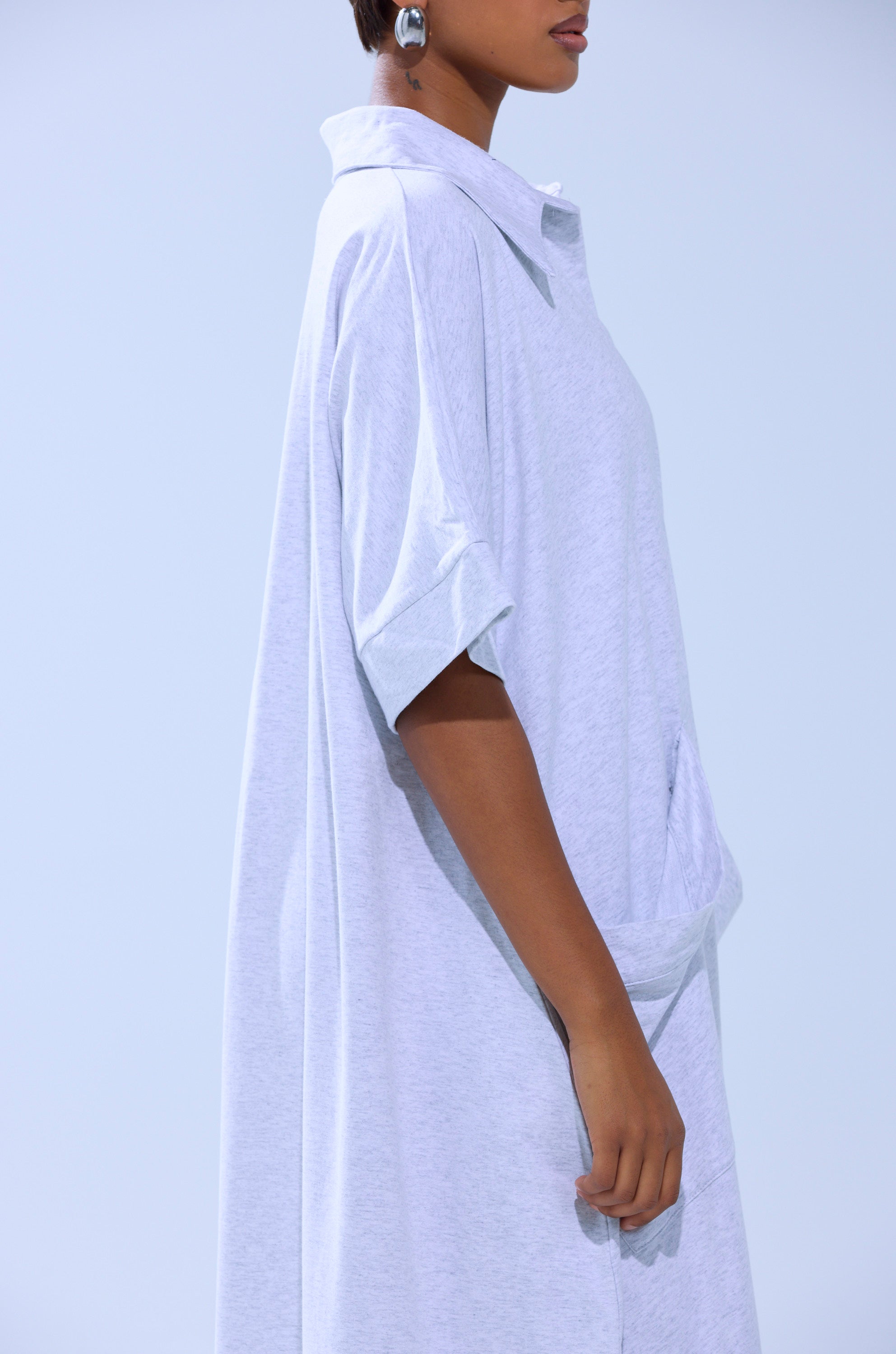 A person in the SHE'S RICH OVERSIZED JUMPSUIT IN HEATHER GREY, featuring short sleeves and a wide collar, stands sideways before a plain light background. They have short hair, large round earrings, and exude understated quiet luxury.