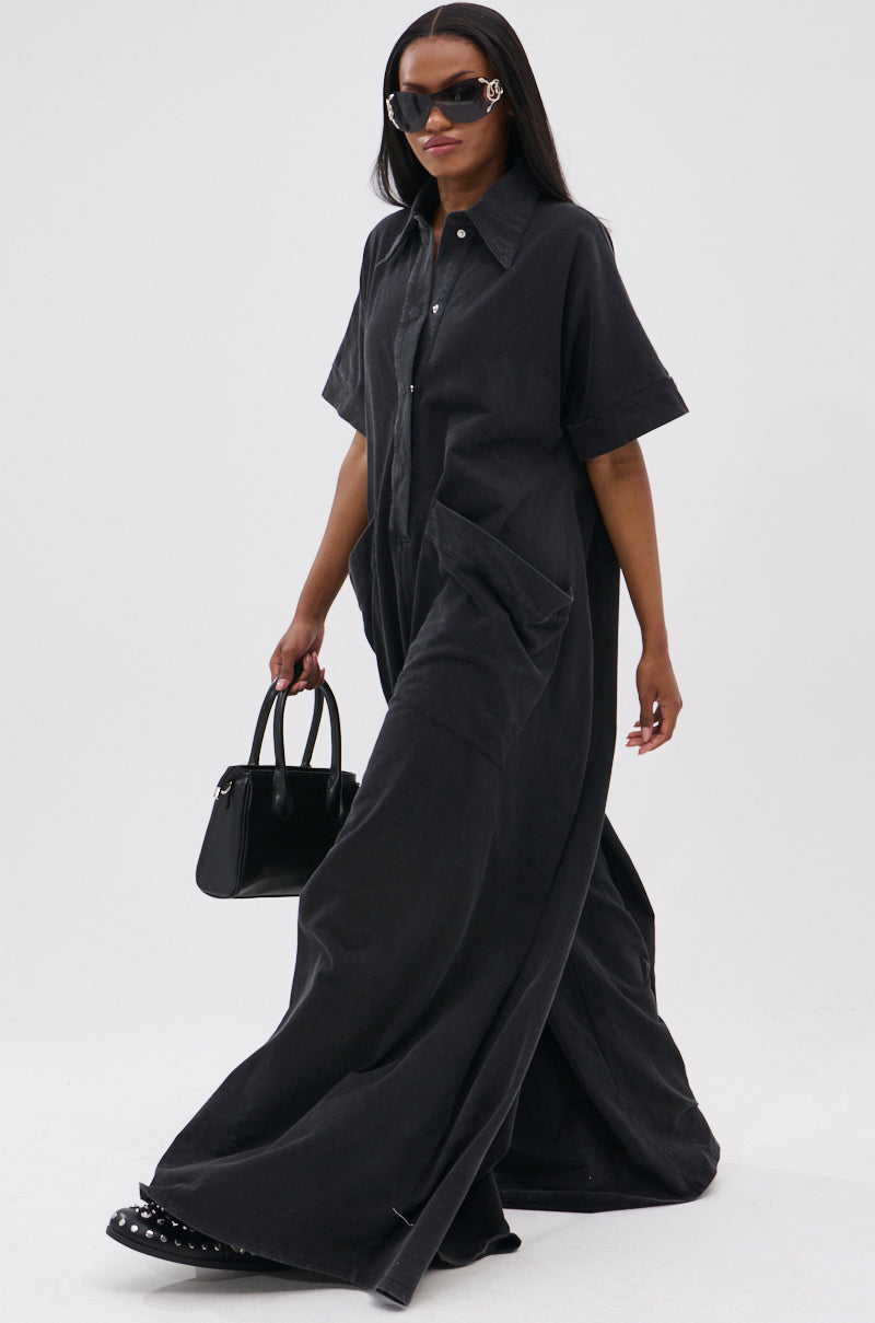 A woman in large black sunglasses walks, carrying a small black handbag and wearing the HAPPY HOUR FRENCH TERRY JUMPSUIT—an oversized, semi-stretchy knit cotton jumpsuit with short sleeves, large front pockets, and black shoes.
