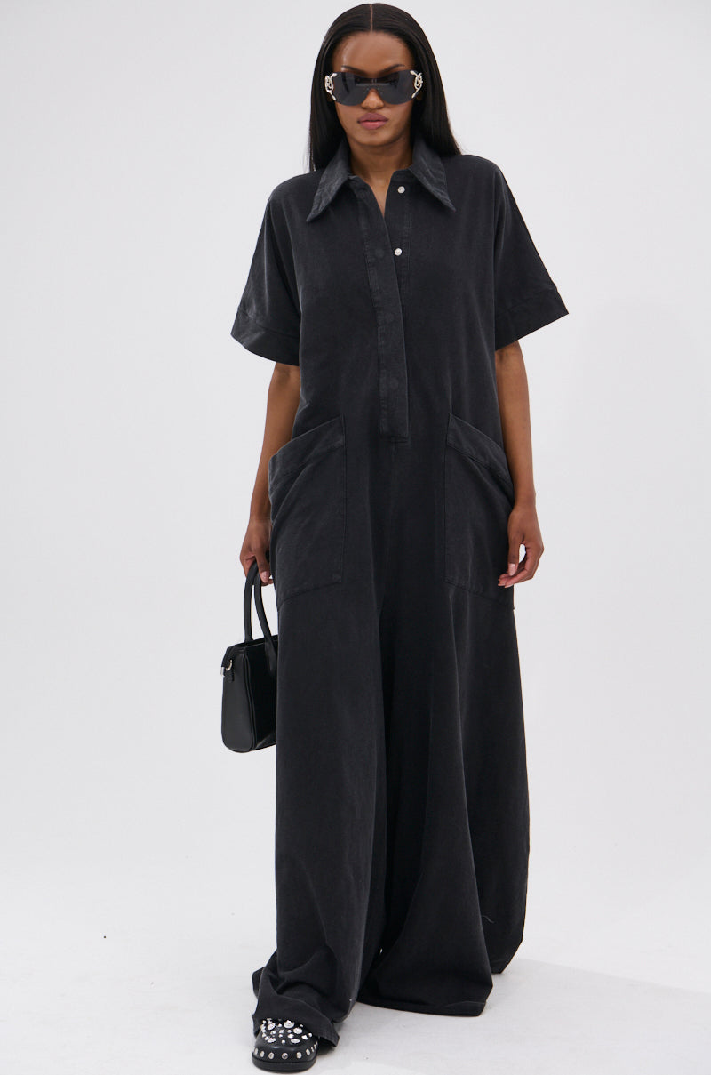 A woman in dark sunglasses wears the HAPPY HOUR FRENCH TERRY JUMPSUIT—loose, floor-length, black, collared with short sleeves and large front pockets—holding a small black handbag against a plain white background.