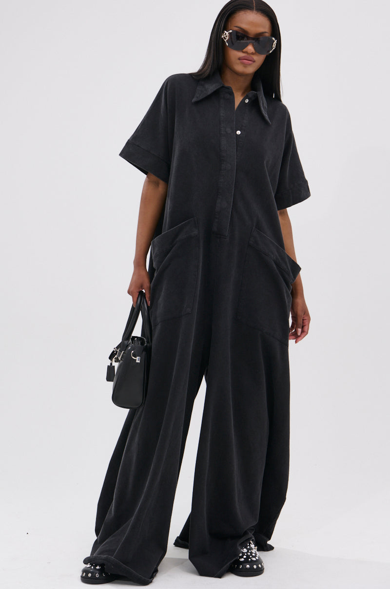 A woman poses in the HAPPY HOUR FRENCH TERRY JUMPSUIT featuring an oversized fit, wide legs, and large front pockets. She pairs it with studded black shoes, sunglasses, and a black handbag against a plain white background.